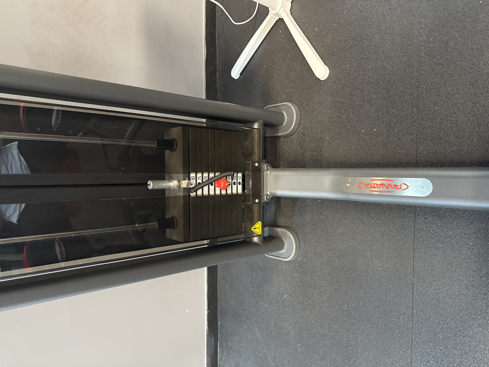 The image shows a close-up, head-on view of the weight stack of a gym machine, likely a cable crossover or lat pulldown machine, in an indoor gym setting. The machine is predominantly grey and black, with a stack of black metal plates visible. A metal rod runs through the center of the weight stack, with a selector pin attached to a red handle at approximately the 40lb mark. Labels with weight increments (10, 15, 20, 25, 30, 40, 45, 50) are visible on a white sticker to the right of the selector pin. A yellow triangular warning sticker with an exclamation mark is attached to the base of the machine. The machine stands on a black rubberized gym floor. To the right of the machine, a white, multi-legged object, possibly a part of another piece of gym equipment or a stand, is visible. The background consists of a light grey wall. The overall environment suggests a commercial gym. The image lacks any indication of the city of Riga, Latvia.