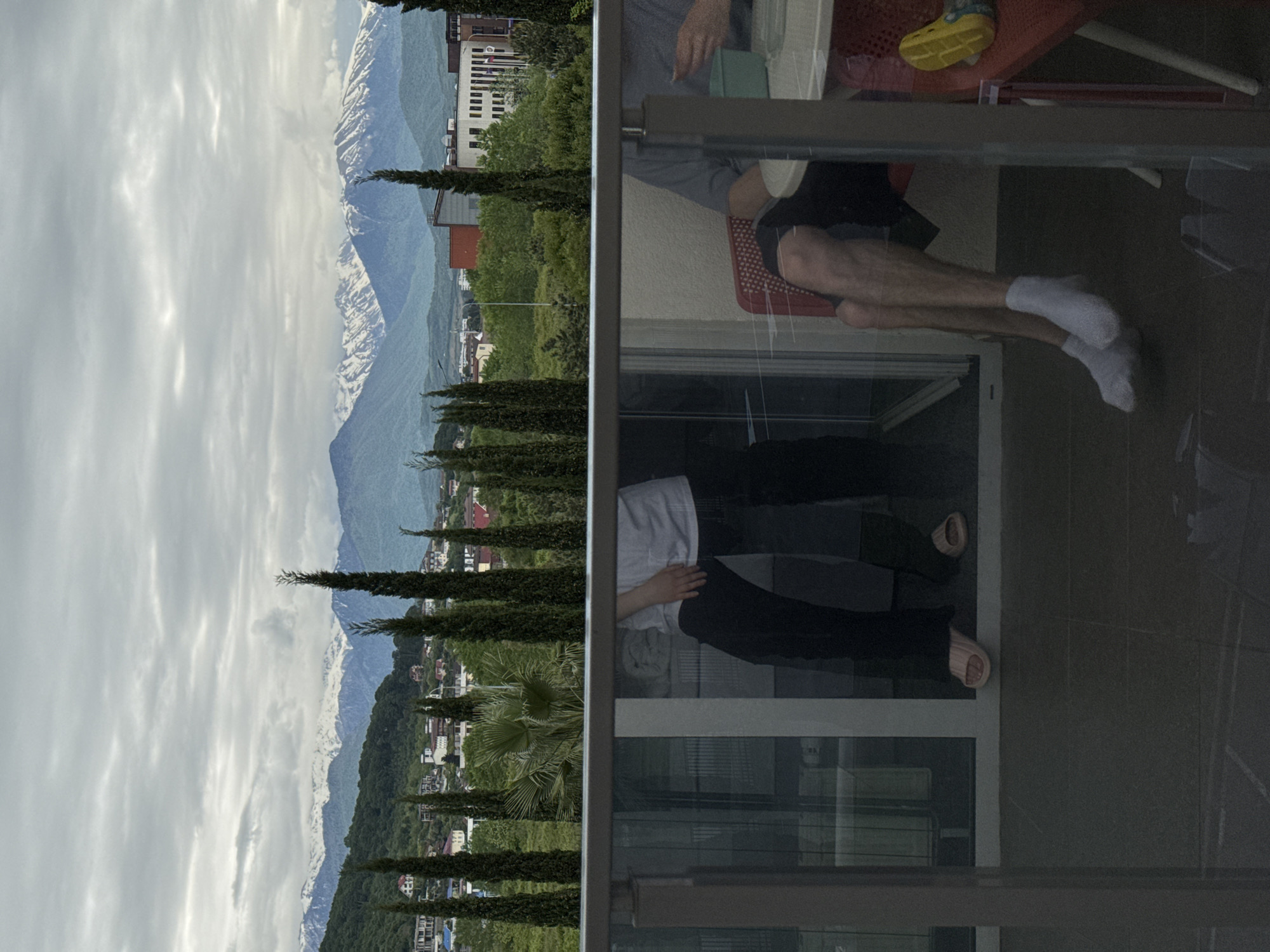 The image shows a view of the city of Sochi, Russia, reflected in a window. The left side of the image shows a landscape with mountains, clouds, and trees. The right side shows the reflection of people and objects within the room, including a person sitting on a red chair with bare feet in white socks, another person standing with a white shirt and black pants, and a yellow sandal on a red object. The image is taken from inside a building looking out through the glass, with the outside scene visible through the reflection.