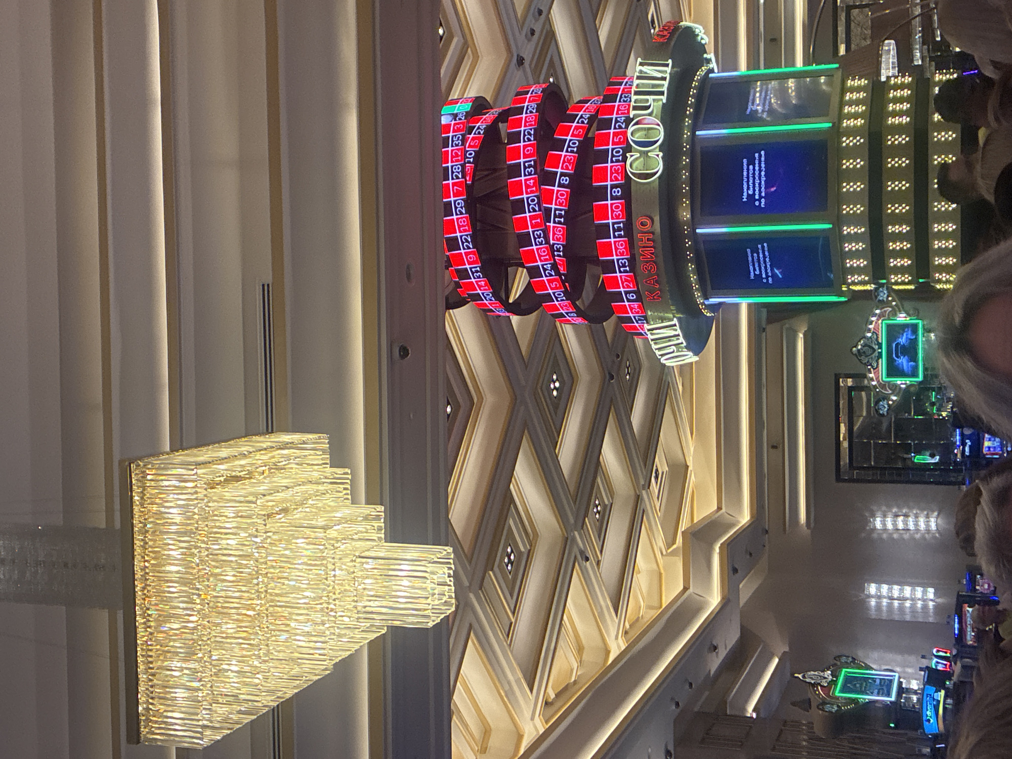 This is a medium shot image of the interior of a casino, likely in Estosadok, Russia, given the "Sochi Casino" signage. The perspective is slightly upward, capturing a grand chandelier on the left and a spiraling display showing roulette numbers on a tall, cylindrical structure in the center. The ceiling is ornate, with geometric patterns and recessed lighting.
In the foreground, partially visible are the heads of several individuals, suggesting they are guests or patrons within the casino. The focus is on the casino's interior design and a prominent roulette wheel display. The roulette display is a series of LED screens wrapped around a central column, showing red and black segments with numbers. Text in Cyrillic script, reading "КАЗИНО" (Casino) and "СОЧИ" (Sochi), is illuminated on the structure below the roulette display. The overall atmosphere appears luxurious and modern, typical of a high-end entertainment venue.