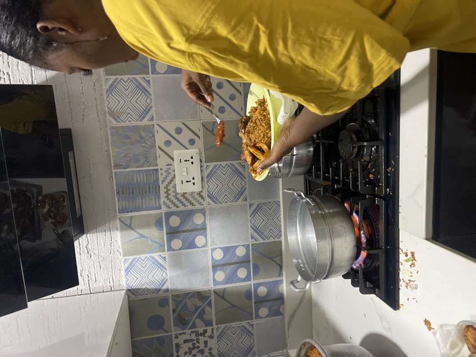 The image depicts a person in a kitchen, engaged in the act of cooking or serving a meal. The individual, visible from the chest up and wearing a yellow t-shirt, is holding a light green plate laden with Jollof rice, fried plantains (dodo), and possibly chicken pieces. Their other hand holds a fork with an orange-colored piece of food, potentially another piece of plantain or a sauce component, positioned over the plate.
The kitchen setting features a gas stove with four burners, where a metal pot is actively heating over a blue and orange flame on the front-right burner. A second metal pot is partially visible in the bottom right corner. The wall behind the stove is adorned with a variety of blue, white, and grey patterned tiles, showcasing geometric, abstract, and subtle floral designs. A white electrical outlet with two sockets is mounted on the tiled wall. A clean white countertop is visible beneath the stove and to the left. Scattered around the gas burners are visible spills or debris, likely rice grains and other food particles, suggesting recent cooking activity.
This scene provides a glimpse into a domestic kitchen, likely within Adenta Municipality, Ghana, where a popular local dish featuring Jollof rice and plantains is being prepared or plated. The lighting indicates an indoor setting. No specific text is clearly decipherable within the image.