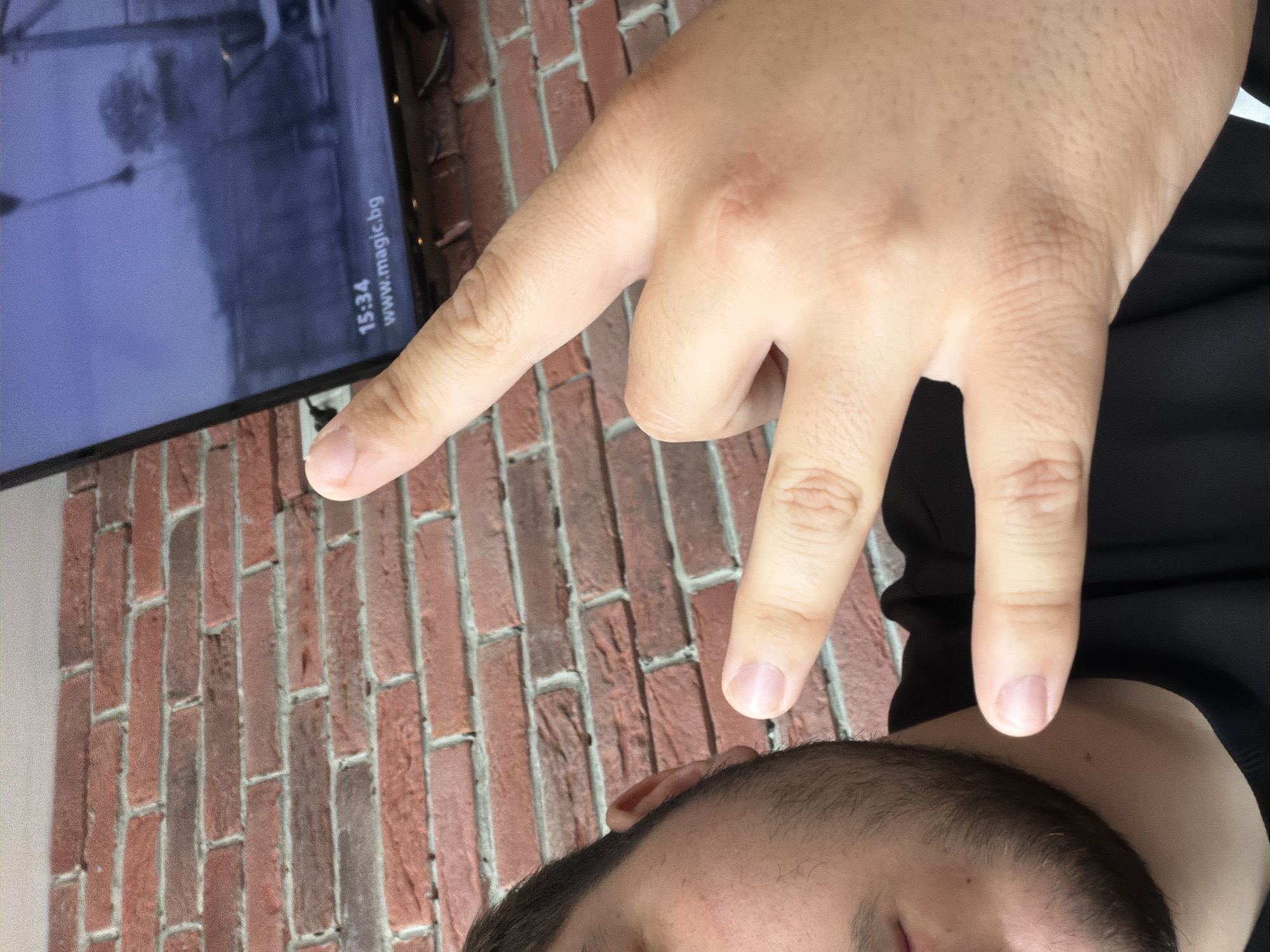 The image, rotated counter-clockwise, depicts an indoor scene with a person's partially visible face and hand in the foreground. The person's head, showing dark hair and a beard, is visible from the top right, while their right hand is held up in the center. Three fingers (index, middle, and ring finger) are extended and spread apart, possibly forming a "W" or "peace" gesture, pointing towards a television screen to the upper left.
The television screen displays a dim, possibly grey or blue-toned, indistinct outdoor scene, potentially security footage or a muted broadcast. Visible in white text on the lower right of the screen are "15:34", indicating the time, and "www.magic.bg", which is a web address.
The background consists of a reddish-brown brick wall with light grey mortar. A sliver of white or light-colored wall is also visible on the far left. The person is wearing black clothing, visible on their shoulder and upper arm. The image captures a casual moment, with the hand gesture seemingly drawing attention to the details on the screen. Given the "magic.bg" text and the provided context, the location is likely Sofia, Bulgaria.