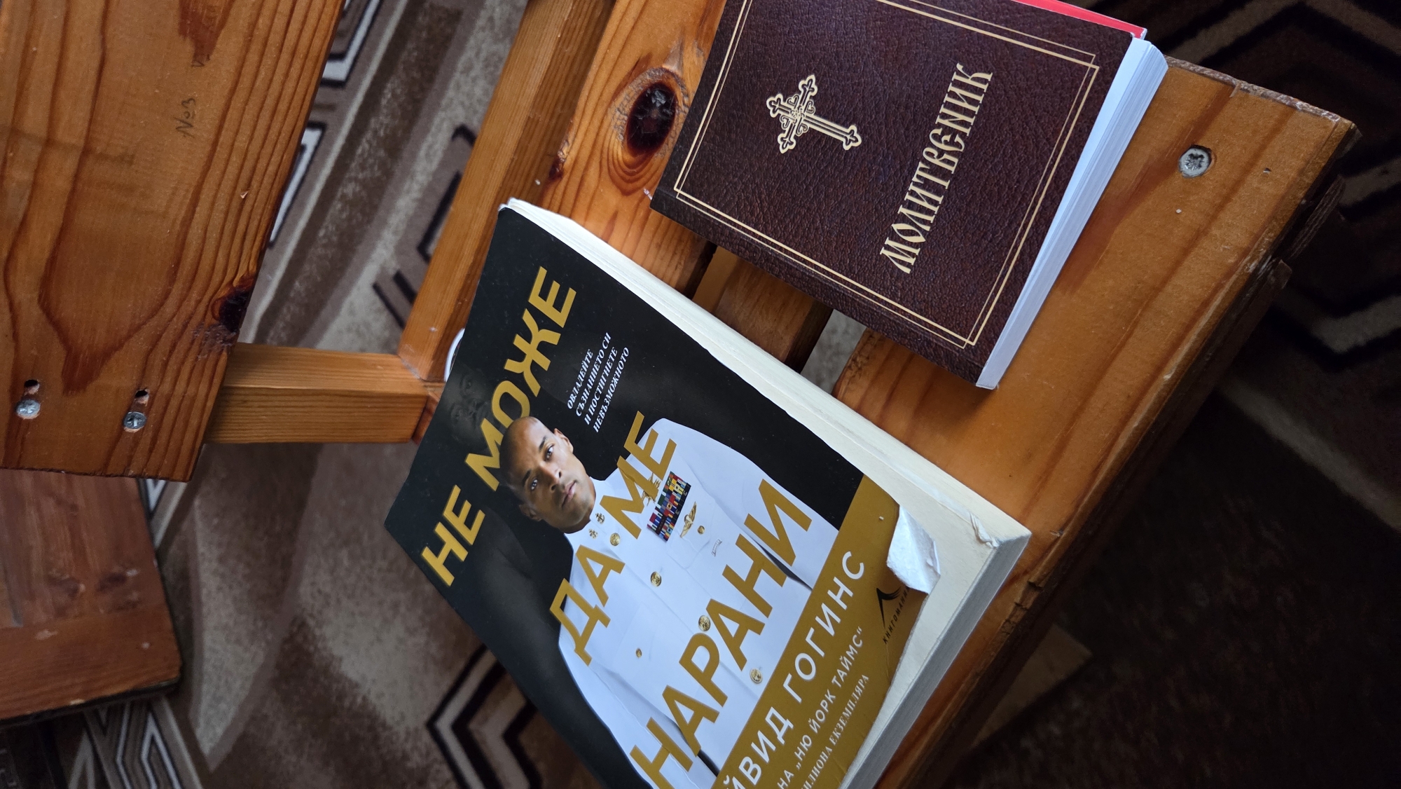 This is a top-down, medium shot photograph taken in an indoor environment, likely a room with wooden furniture. The main subjects are two books lying on a wooden surface.
The book on the left is a large hardcover with a black cover featuring a picture of a man in a white military uniform. Large yellow text overlays the image. The text appears to be in Cyrillic script. The book is titled "НЕ МОЖЕ ДА МЕ НАРАНИ" which translates to "It Can't Hurt Me." Beneath the main title, there is smaller text that seems to be author attribution and promotional material, including "ЗАВИСНОСТИ", "ЗАВИСНОСТИ", and "НЮ ЙОРК ТАЙМС". A torn piece of a yellow book jacket is visible on the bottom edge of this book.
The book on the right is smaller and bound in dark brown faux leather. It has a gold embossed border and a gold cross symbol on the cover. The title, also in Cyrillic script, reads "МОЛИТВЕНИК," meaning "Prayer Book."
The wooden surface is a slatted bench or chair, showing the grain of the wood and some screws. A rug with a patterned design is visible in the background. The lighting is moderate, casting soft shadows. There are no people or animals present in the image.