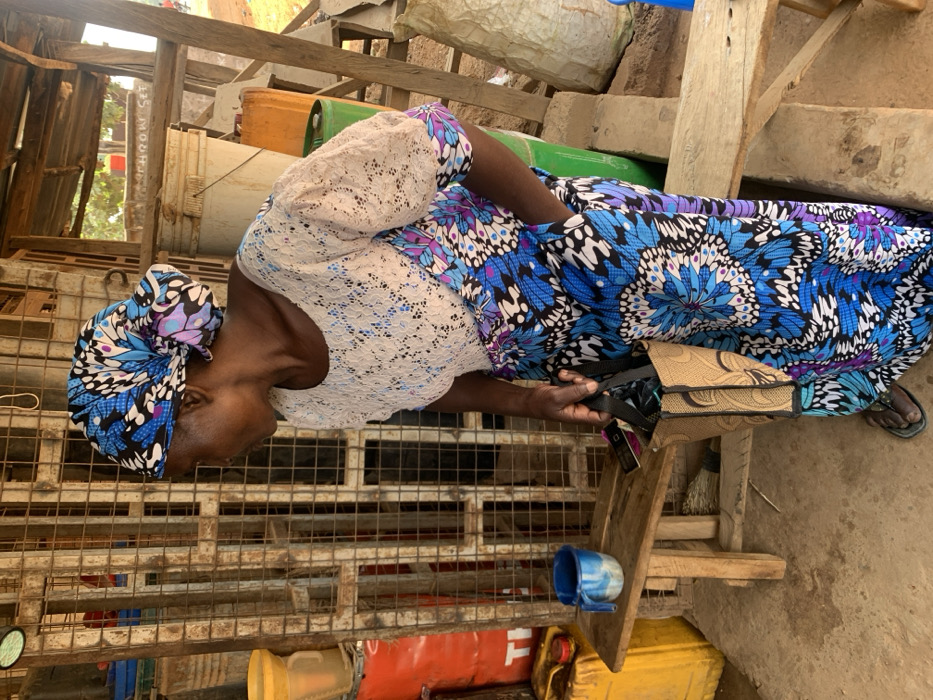 An adult female is depicted standing in a semi-outdoor commercial setting in Ube, Nigeria. She wears a blue, black, and white patterned headwrap and a matching long skirt or dress, combined with a white-cream lace short-sleeved top. Her right hand holds a small, light brown woven bag with black handles. She wears dark flip-flops. Her gaze is directed downwards and slightly to her left. The immediate background features a rusted metal grate structure. To her left, a yellow plastic funnel rests atop a partially visible red metal barrel marked "TI". A small, light brown wooden bench with a blue and white speckled mug is situated at floor level to her left. To her right, a large white plastic bucket, an orange plastic jerrycan, and a green plastic jerrycan are visible. The scene is structured by rudimentary wooden posts and beams, and the ground is an uneven, dirt-colored surface.