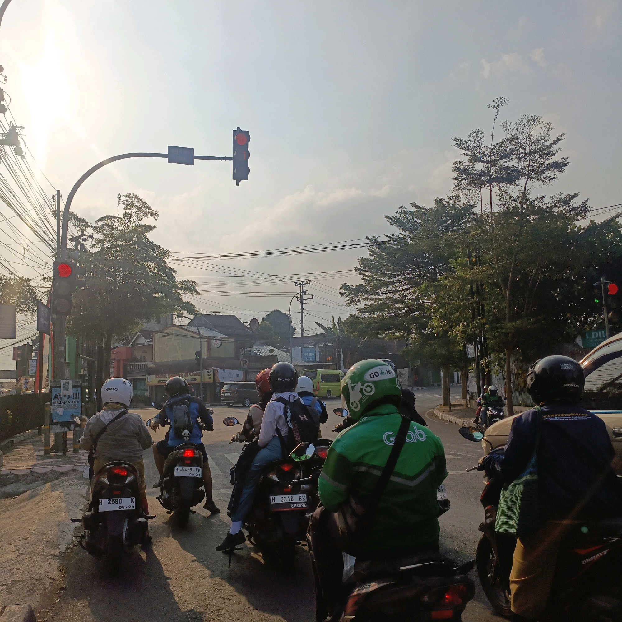 The image depicts a street scene in Salatiga, Indonesia, under a bright, hazy sky, likely in the late afternoon given the warm lighting and long shadows. Several motorcyclists are stopped at a traffic light which is red. The foreground is dominated by the backs of several motorcyclists and their bikes. The rider in the center foreground wears a white helmet and a light gray jacket over a white shirt and a patterned scarf. They are seated on a dark motorcycle with a license plate reading "H 2590 K" and "08-28". To their right, another motorcyclist wears a blue jacket over a dark shirt and a dark backpack, their motorcycle's license plate reads "H 5428 VK" and "06-31". In the right foreground, a rider in a bright green GoJek jacket is partially visible, facing away from the camera.
Further into the scene, other motorcyclists are visible. One individual wears a white helmet and a dark backpack, and another in a green helmet also appears to be a GoJek rider. A small green van is visible in the middle ground. The street is lined with trees and buildings, some of which are painted in various colors. Electrical wires are strung across the sky, common in urban Indonesian landscapes. The traffic light in the upper left corner is displaying a red signal, and another red light is visible on a traffic signal further down the street to the right. The overall atmosphere suggests a typical urban traffic situation in Indonesia.