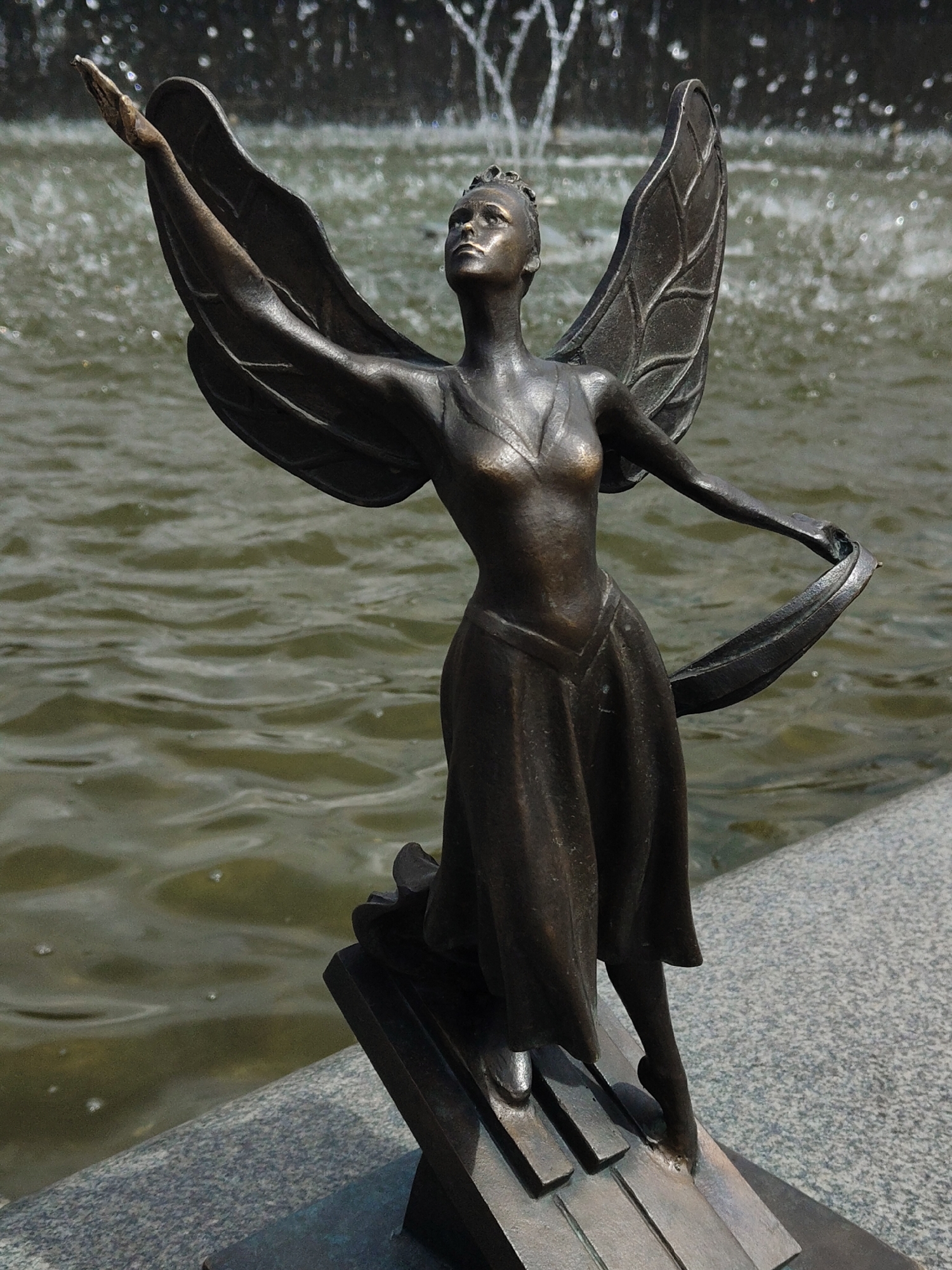 The image displays a close-up of a bronze statue depicting a graceful female figure with wings, set against a backdrop of rippling water from a fountain. The statue's figure looks upwards with an aspirational gaze. Her left arm is extended and slightly raised, while her right arm is bent, holding a piece of flowing fabric that drapes towards her side. She wears a flowing dress with a V-neckline and has bare feet. One foot is sculpted to appear as if on pointe, resting on a base designed to resemble the keys of a piano. The wings are detailed with a leaf-like vein pattern. A delicate crown or headpiece adorns her head.
The background features dark, murky green water with visible ripples and splashes from a fountain, suggesting movement and activity. The statue is positioned on a speckled grey granite or stone ledge that separates it from the water. The scene is brightly lit, indicating a clear, sunny daytime. There is no visible text on the statue or in its immediate surroundings. The overall impression is one of artistry, music, and inspiration, perhaps representing a muse or a dancer.
