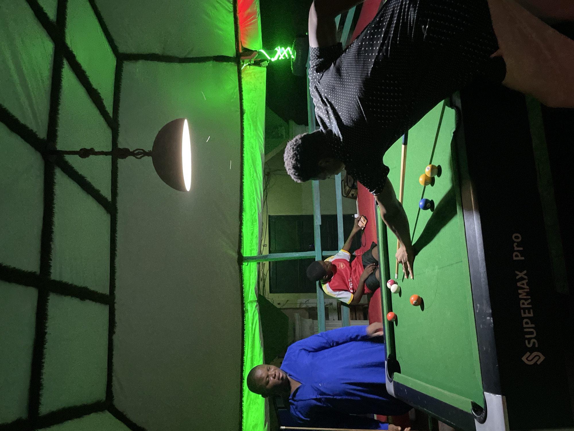 This image captures an indoor recreational setting in Lokoja, Nigeria, under low-light conditions dominated by artificial green illumination. A game of billiards is taking place on a 'SUPERMAX Pro' branded pool table.
Three males are present in the scene. To the left, an adult male in a blue tunic-style garment stands, observing the pool table. In the center-background, a younger male wearing a red and white football jersey and dark shorts is seated on a red upholstered bench, engaged with a smartphone. In the foreground, an adult male, viewed from behind, wears a dark, polka-dotted short-sleeved shirt and light-colored trousers, leaning over the pool table to execute a shot with a pool cue.
On the green felt playing surface are a white cue ball and several object balls: a solid blue ball (numbered 2), two solid yellow balls (numbered 1 and 9), a solid orange ball (numbered 5), a solid red ball (numbered 7), and one red-and-white striped ball. A single, metallic, dome-shaped lamp hangs directly over the table, providing focused bright white light downwards. The ceiling and upper wall perimeter are covered in a light-colored, textured material, sectioned by dark grid lines, intensely illuminated by green light strips installed along the upper edge of the structure. In the background, behind a low wooden railing, details of a building structure with dark window or door frames are visible. A distant green light fixture emitting a zig-zag pattern is also discernible on the far right.