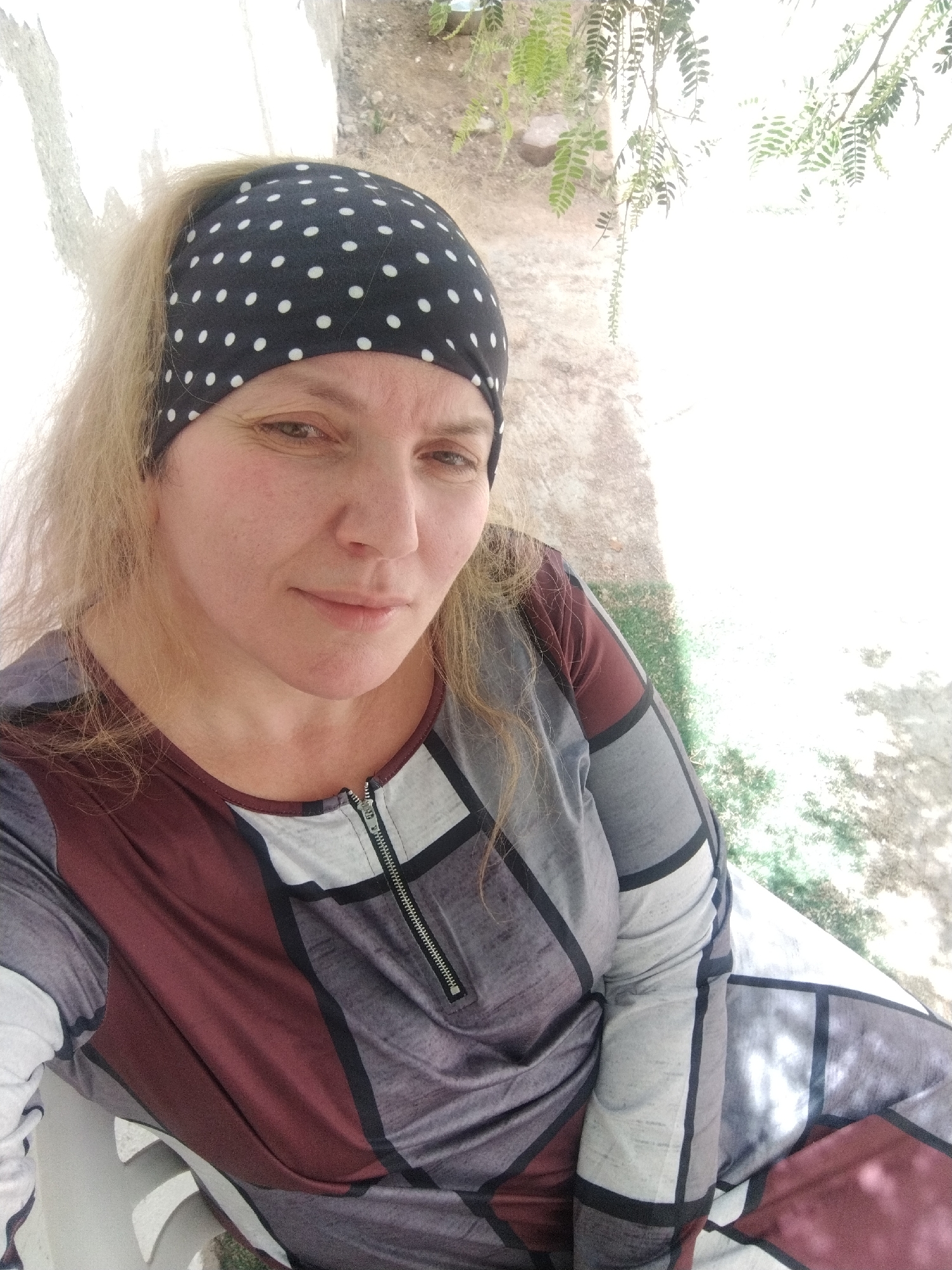 A woman with blonde hair and a polka-dot headscarf is seated outdoors. She is wearing a long-sleeved shirt with a geometric pattern and a zipper detail near the collar. The location is Al Mas'oudi, United Arab Emirates, evident by the background vegetation and setting. The woman is looking at the camera, with a neutral expression. She is sitting on a white chair or bench. The setting appears to be a garden or outdoor area, with a whitewashed wall and some greenery visible. The lighting is bright.