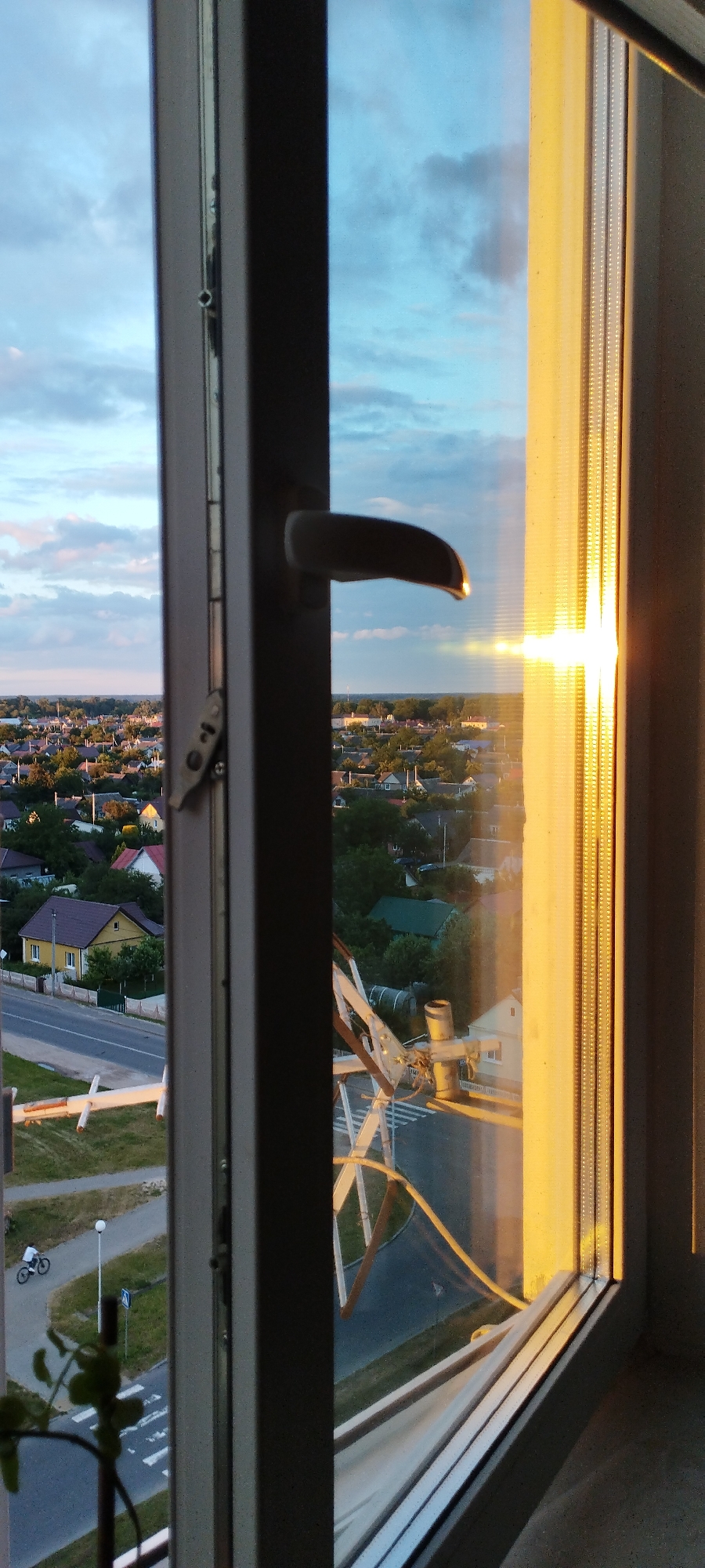 The image shows a view through a window frame looking out at a suburban landscape during what appears to be sunset. The sun is setting to the right, casting a bright, warm glow on the right side of the window frame and reflecting off the window pane. In the foreground of the window, a dark handle is visible, suggesting the window is partially open.
The landscape outside consists of numerous houses with pitched roofs, surrounded by trees and green vegetation. A road is visible in the middle ground, with a pedestrian crossing and a street lamp. A person on a bicycle is riding on the road. In the distance, the sky is a mix of blue and clouds, with the sunset colors becoming more pronounced.
The location is identified as Skidzieĺ, Belarus, which is consistent with the observed suburban architecture and vegetation. The overall scene captures a peaceful, everyday moment in a residential area as the day concludes.