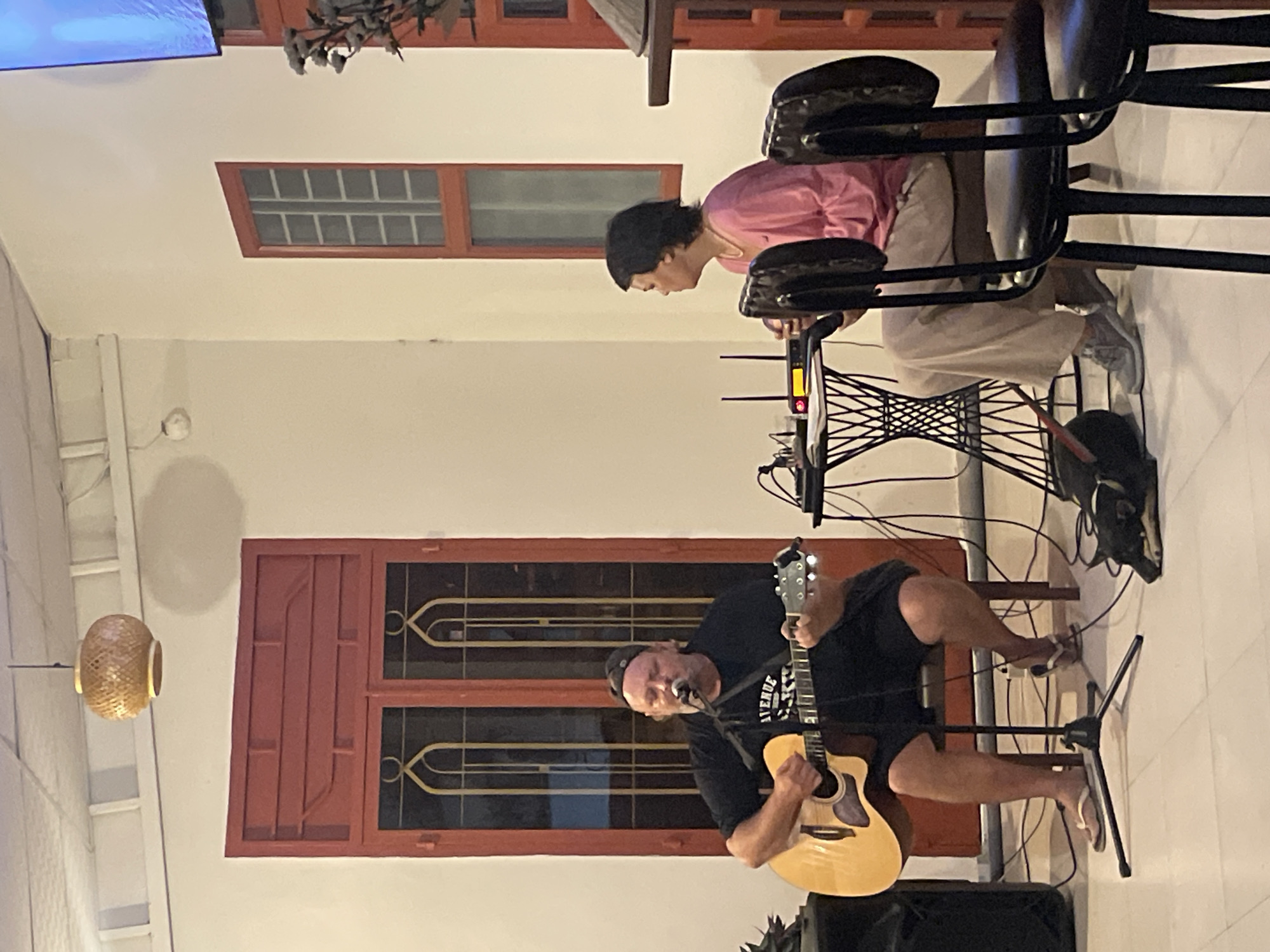 This image captures an indoor, evening scene in what appears to be Bac Natural Heritage Area Trang, Vietnam, featuring a casual musical performance.
In the foreground, a man is seated, passionately playing an acoustic guitar and singing into a microphone. He has a beard, wears a dark t-shirt with "AVENUE" visible text, shorts, a baseball cap worn backward, and white flip-flops. He is focused on his performance, his mouth open as if singing. An amplifier or speaker rests on the floor near his feet.
Further back, to the right of the performer, a woman is seated on a tall stool. She has short dark hair and is wearing a pink long-sleeved top and light-colored trousers. She leans forward, looking down at electronic equipment, possibly a router or modem, placed on a table. This table appears to be an antique sewing machine base repurposed. Cables are visible around the equipment and leading to the performance area.
The room has light-colored walls and features two prominent windows with reddish-brown frames and decorative grates. One window is above the performer, and another is behind the seated woman. A traditional-looking bamboo light fixture hangs on the left side of the frame. In the upper left corner, a portion of a television screen displaying a blue image is visible. The overall setting suggests a comfortable, intimate gathering or event.