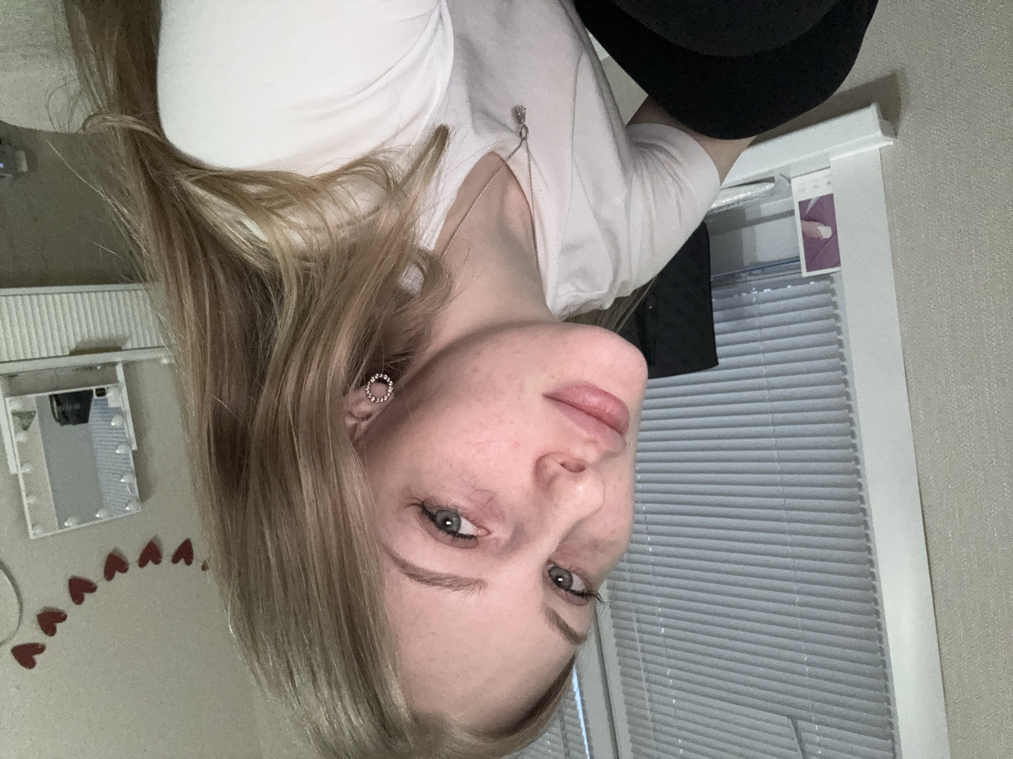 The image displays a close-up portrait of a young woman, presented rotated 90 degrees clockwise. She has fair skin with visible freckles across her nose and cheeks, light blue or grey eyes, and long, straight to slightly wavy blonde or light brown hair. Her expression is neutral, and she looks directly at the camera. She is wearing a white t-shirt and a simple silver chain necklace with a small, circular pendant. A hoop earring, adorned with small, clear stones, is visible on her left ear.
The background indicates an indoor setting. To what would be her left if the image were upright, a rectangular vanity mirror with visible light bulbs around its frame is mounted on a light-colored wall. Below the mirror, a curving line of small, red, heart-shaped decorations is affixed to the wall. To her right, a window covered by horizontal blinds allows natural light into the room. A small, purple-toned card or photo is tucked into the top frame of the window blinds. The lighting is soft and even, suggesting a daytime scene. No legible text is visible in the image. The provided location context is Perm, Russia.