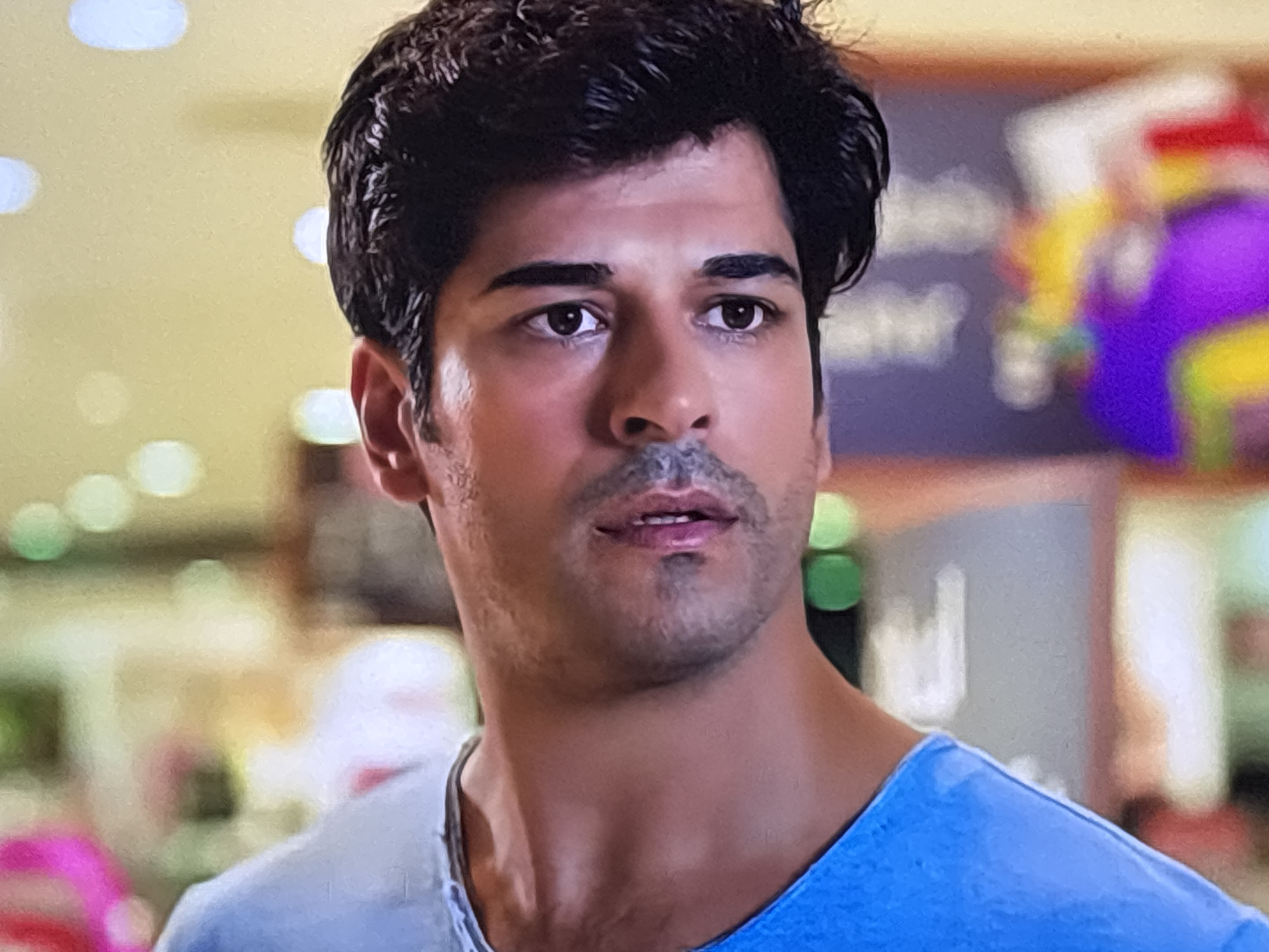 A close-up shot of a man with dark hair and brown eyes, wearing a blue t-shirt. His expression is neutral. The background is blurred, revealing hints of a retail environment with lights and colors, suggesting he is indoors in a public space. There is no information regarding Sofia, Bulgaria.