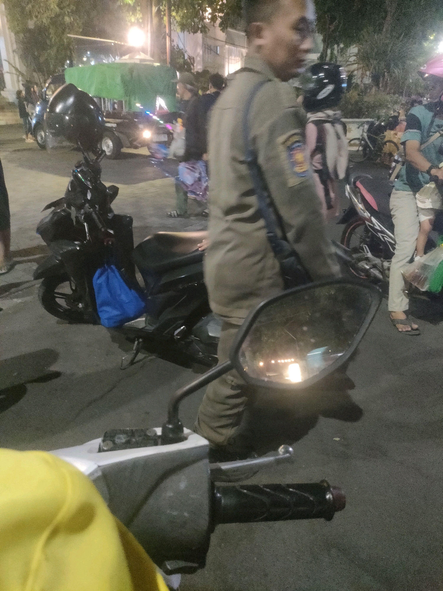 In Surabaya, Indonesia, at night, a person in a khaki uniform is captured from the chest down, walking past parked motorcycles. One motorcycle, a black scooter, is in the foreground with a bright blue bag hanging from its side. Behind it, another motorcycle, possibly a pink and black model, is parked. Further in the background, a flatbed truck covered with a green tarp is visible. Several other individuals are present in the scene, some partially obscured. The environment appears to be an outdoor, urban setting with artificial lighting contributing to the dimly lit atmosphere.