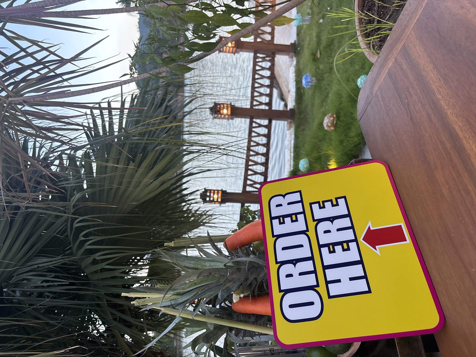 An "ORDER HERE" sign with an arrow pointing down is placed on a wooden table in the foreground. Behind the sign, on the table, are pineapples and carrots. The background shows a body of water, a wooden fence with lamps, lush green grass, and palm trees. The scene appears to be outdoors, possibly at a restaurant or a food stall.