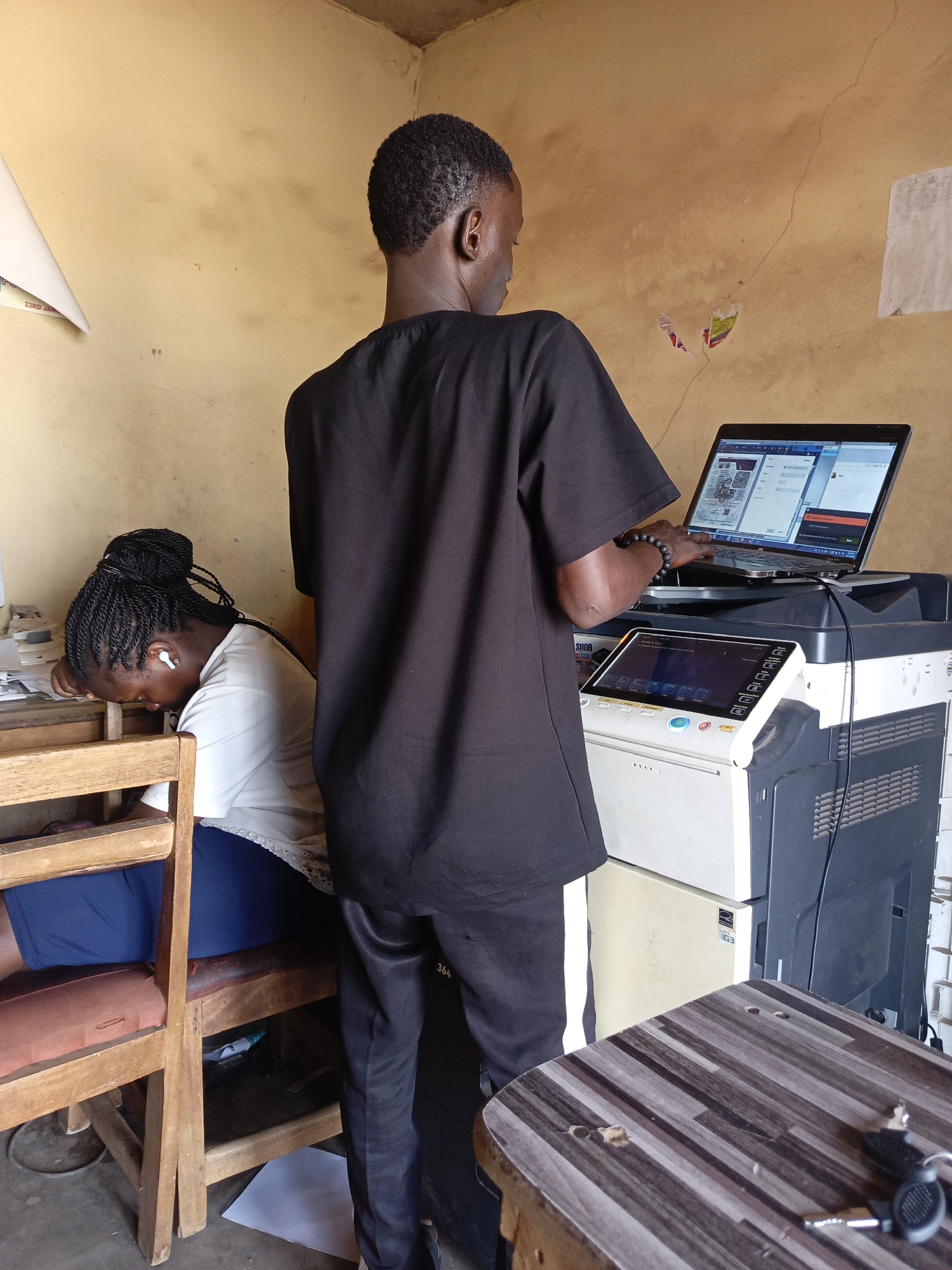 The image captures an indoor scene in what appears to be a small office or business, likely in Akwanga, Nigeria. The setting is characterized by plain, light brown/yellow walls showing signs of wear, including stains and cracks.
In the foreground, a young man, seen from behind, stands facing a large multifunction printer/copier. He wears a black t-shirt and dark trousers, with a beaded bracelet on his left wrist. He is actively using a laptop, which is placed on top of the printer. The laptop screen displays what looks like multiple open applications or browser tabs, including a document with images and text, suggesting he is engaged in computer-related work, possibly involving printing or scanning.
To the man's left and slightly in the background, a young woman is seated at a wooden desk or table, her back partially visible. She wears a white t-shirt, blue clothing on her lower half, and has braided hair pulled back. She is leaning forward with her head down, appearing to be resting or sleeping, with white earbuds in her ears. The wooden chair she sits on is simple, with a padded seat.
Other objects in the scene include a small wooden table with a patterned surface in the bottom right, where a set of keys is placed. Various papers are visible around the woman's desk area and beneath the man's workstation. A white triangular object, possibly part of a sign or display, is affixed to the wall near the top left, with partially visible text "23RD JANU" and "SUDA". The large printer/copier has a digital display showing various icons and text, and an "ENERGY STAR SUPER G3" label is visible on its side. A number "364" is faintly visible on the wall near the floor. The overall impression is that of a functional but modestly equipped local service point during daytime hours.