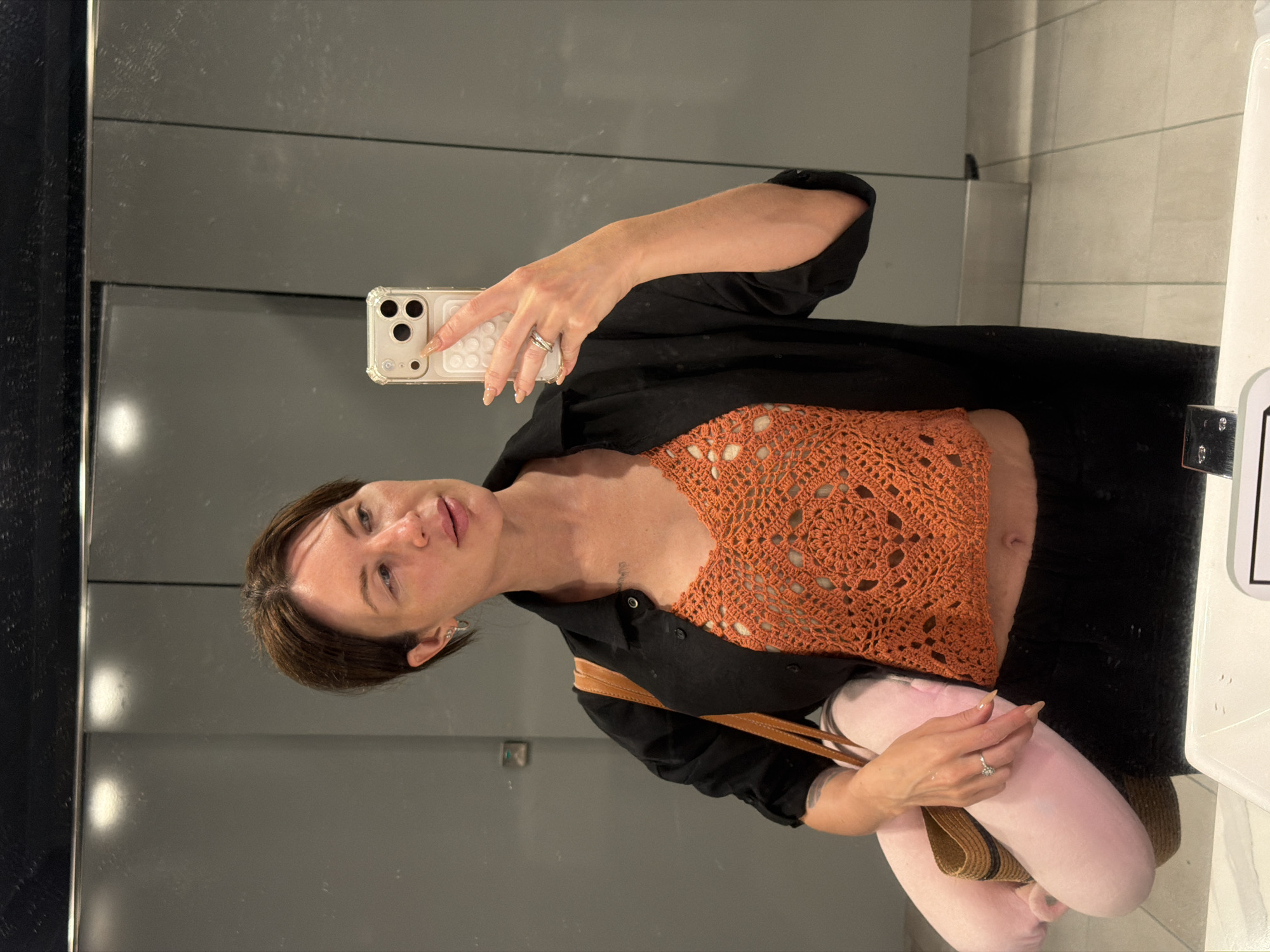 A woman with short brown hair takes a mirror selfie in a public restroom. She is wearing a black unbuttoned shirt over an orange crocheted crop top and black pants. She has a tan crossbody bag with a pink neck pillow slung over her shoulder. She is wearing silver hoop earrings and a silver ring on her left hand. The restroom has gray tiled walls and a white countertop in the foreground.