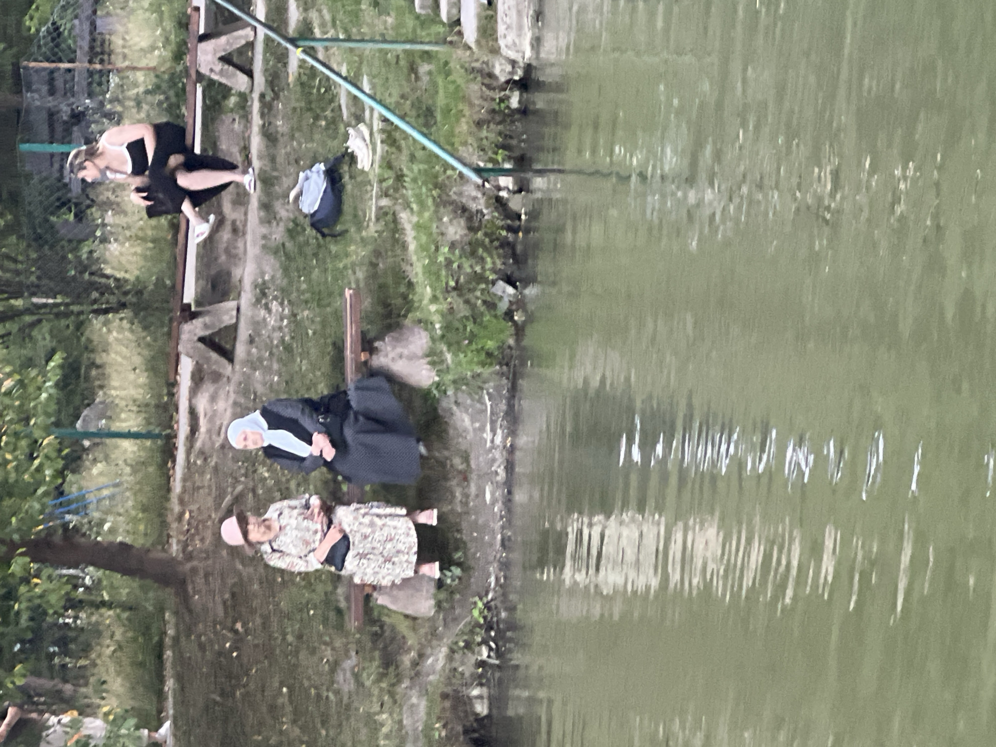 Three women are seated on a bench near a body of water. Two women are on the left side of the bench, and one woman is on the right. The woman in the center on the left is wearing a floral dress and a pink hat. The woman to her left is wearing a black dress and a light blue hijab. The woman on the right is wearing a black dress with white trim and is looking to her left. The bench is made of wood and is supported by concrete blocks. Behind the bench is a grassy area with trees and a fence. To the right of the bench is a set of stairs leading down to the water. There is a backpack and a pair of shoes on the ground near the bench. The water in the foreground is a murky green color and has ripples on its surface, reflecting the light. The scene appears to be a public park or recreational area.