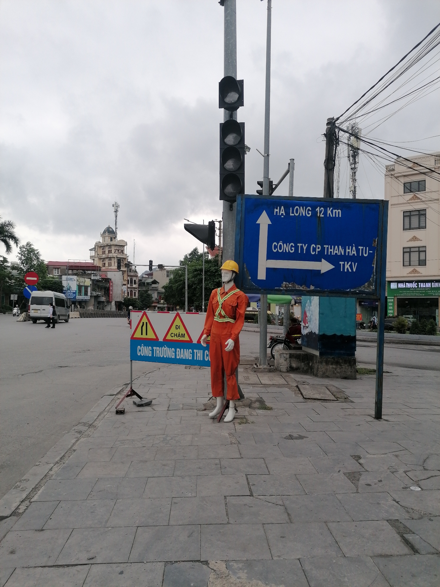 The image depicts an urban street corner or intersection in Ha Tu Ward, Vietnam, under an overcast sky during daytime. In the immediate foreground, on a paved sidewalk, a mannequin dressed as a construction worker stands. It wears an orange jumpsuit with reflective stripes and a yellow hard hat.
Beside the mannequin, a temporary A-frame sign with yellow and red triangles warns of ongoing work. To the mannequin's right, a prominent blue directional sign points towards "HẠ LONG 12 Km" (Ha Long 12 Km) with an upward arrow and "CÔNG TY CP THAN HÀ TU-TKV" (Ha Tu Coal Joint Stock Company - TKV) with a rightward arrow. Traffic lights are mounted on poles next to this sign.
In the background, a bustling street features various buildings, including a distinctive multi-story structure with a domed roof and a telecommunications tower on top. Trees and other urban foliage are also visible. A white minivan and a pedestrian are seen on the road. Numerous overhead utility cables are tangled across the scene. The sidewalk consists of grey square paving slabs, some of which appear worn.
Visible text includes:
* On the construction sign: "ĐI CHẬM" (Slow Down) and "CÔNG TRƯỜNG ĐANG THI CÔNG" (Construction Site in Progress).
* On the blue directional sign: "HẠ LONG 12 Km" and "CÔNG TY CP THAN HÀ TU-TKV".
* On building signs: "DISMY", and partially visible "CHĂM SÓC SỨC KHỎE" (Health Care) and "NHÀ THUỐC THANH BÌNH" (Thanh Binh Pharmacy).