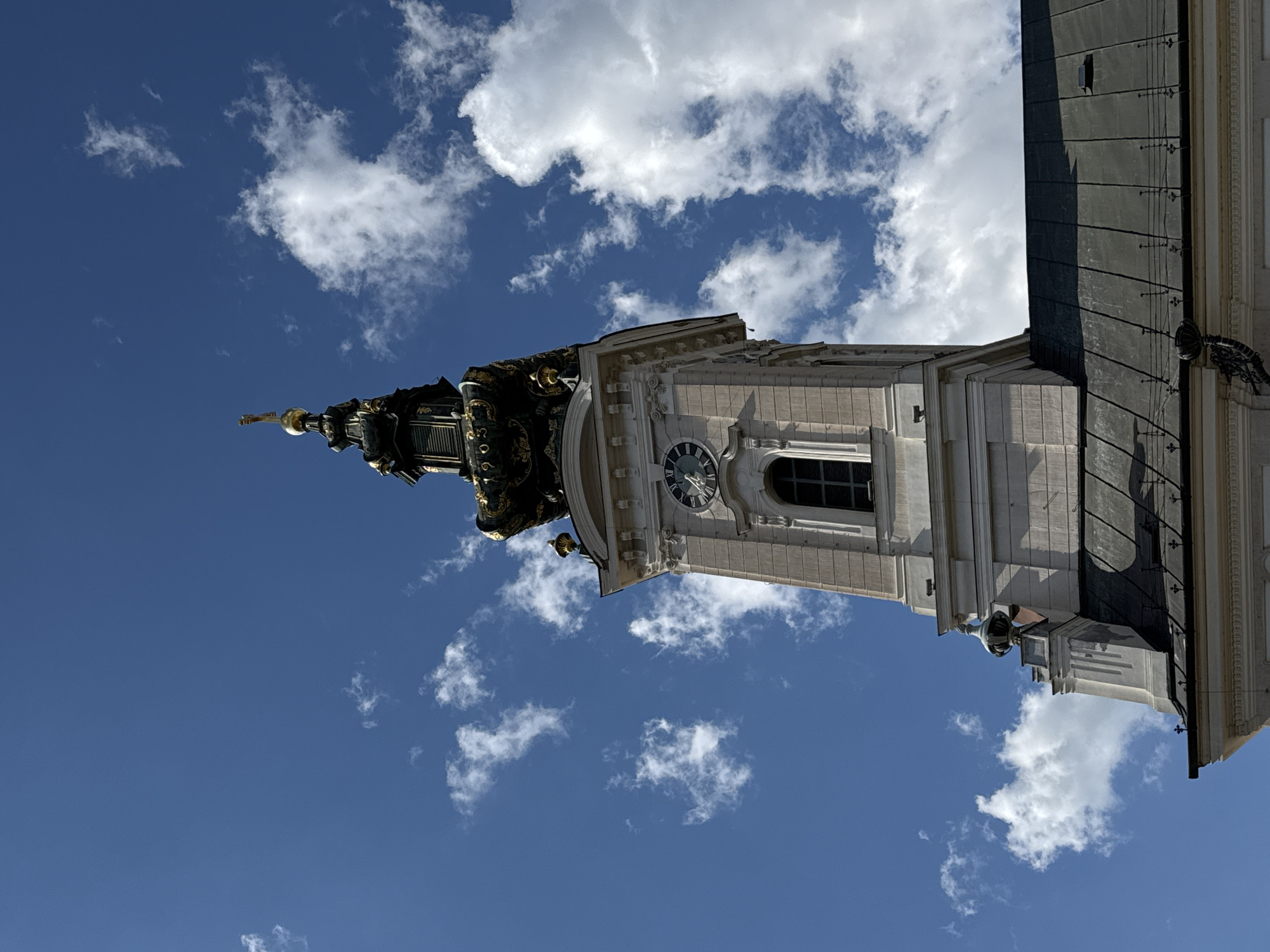 This image displays the upper section of a prominent church tower against a vibrant blue sky with scattered white cumulus clouds. The structure is the Name of Mary Church (Katedrala Imena Marijinog), a Roman Catholic church located on Trg slobode (Freedom Square) in Novi Sad, Serbia.
The central subject is the light-colored stone or stucco tower, which features a circular clock with a dark face and white Roman numerals, indicating a time approximately between 10:10 and 10:15. Below the clock is a recessed, rectangular window fitted with dark louvers or shutters. Ornate architectural details, including cornices and moldings, adorn the tower.
Crowning the tower is an elaborate, multi-tiered spire. The lower part of the spire is dark with visible gilded ornamentation, and the year "1903" is inscribed on one of its dark sections. The spire tapers upwards, culminating in a golden orb topped by a small, golden cross.
To the right of the tower, a portion of the church's dark, possibly metal, roof is visible, sloping downwards. Strong, direct sunlight illuminates the scene, casting subtle shadows on the left side of the tower and highlighting the architectural details. No people or other active subjects are present in the frame.