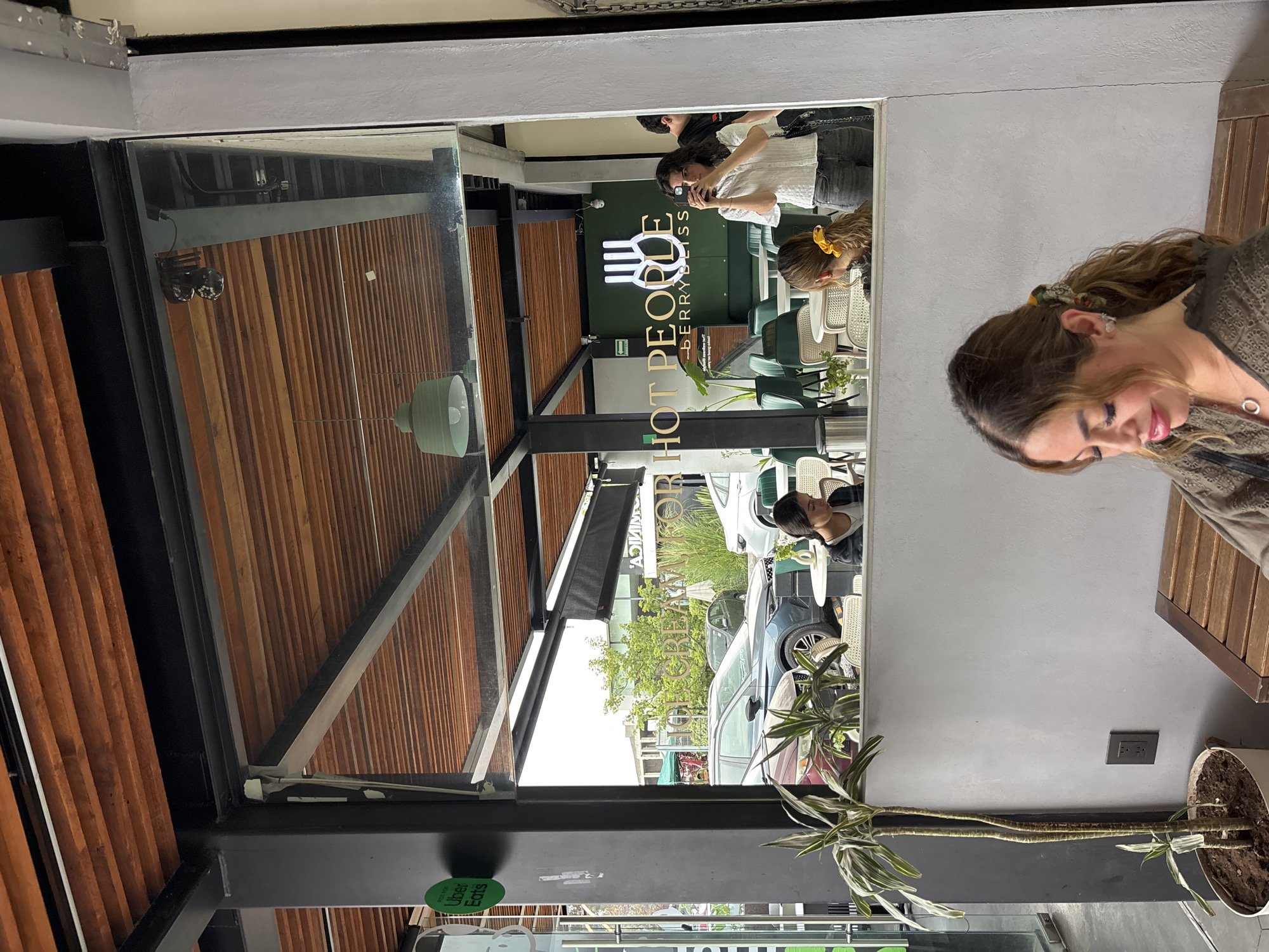 The photo appears to be taken in a cafe in San Bernardino Tlaxcalancingo, Mexico, as it contains the sign for "Hot People Berry Bliss," a cafe with locations there. The image is shot through a glass pane, likely a window or door, reflecting the cafe's interior and exterior. Inside the cafe, one woman is seen taking a photo with her phone. In front of the glass, a woman sits at a wooden table, looking down. Beyond the cafe, a street scene is visible with cars and a few pedestrians. The cafe has a modern industrial design with a wood ceiling. A potted plant sits near the lower edge of the glass, seemingly inside the cafe. A dark, industrial-looking pole with an electrical outlet is on the ceiling adjacent to the glass partition. There are Uber Eats logos visible.