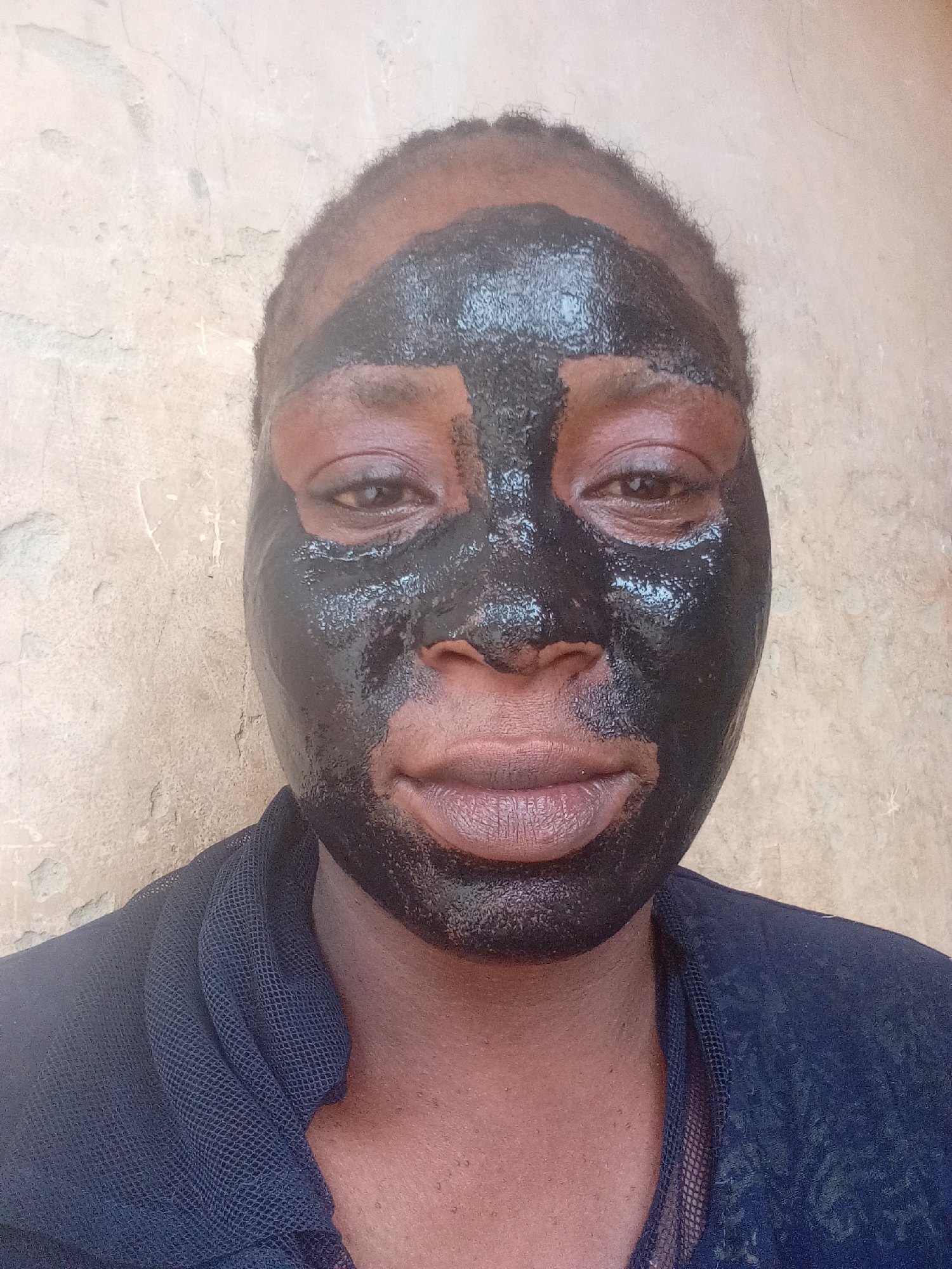 A close-up photo shows a person's face covered with a black facial mask, set against a textured, off-white wall. The person's hair appears dark and cropped close to the scalp. The mask covers most of the face, excluding the lips and small areas around the eyes. The person is wearing a dark, patterned, mesh-like garment. The background wall has a cracked and uneven surface. The location is Akwanga, Nigeria.
