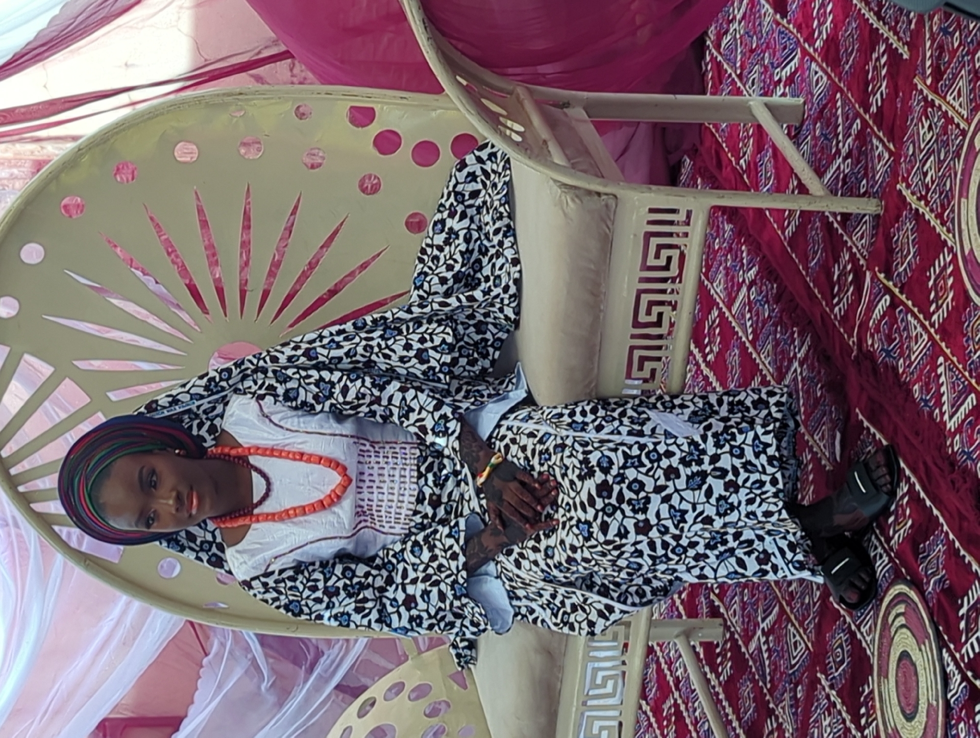 This image, captured in Unguwar Rimi, Nigeria, features a woman seated in a decorated setting, likely for a cultural event or ceremony. The central subject is an African woman dressed in traditional attire. She wears a white top with a patterned front, covered by a flowing garment adorned with black, white, blue, and reddish-brown floral or leaf patterns. Her head is wrapped in a colorful textile showcasing blue, green, and orange stripes. A prominent orange coral bead necklace graces her neck. Her hands, resting in her lap, display dark, intricate designs, possibly henna, and a multi-colored bracelet on her right wrist. She wears black sandals.
She is seated on a light-colored, possibly off-white or beige, ornate chair with a distinctive fan-like cut-out design on its backrest and geometric patterns on its armrest. The background is adorned with draped fabrics, including sheer white and solid pink/magenta materials, creating a festive atmosphere. The floor is covered by a richly patterned red rug with intricate white and blue geometric designs. A circular, woven mat or tray, also patterned, is visible on the rug to the right. The bright lighting suggests it is daytime. The woman maintains a calm and composed expression, appearing to pose for the photograph. No visible text is present in the image.