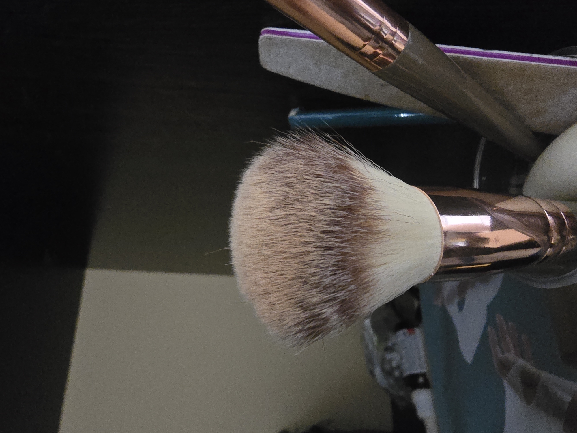 The image shows a close-up of a fluffy makeup brush with a rose gold handle, standing upright in a container. The brush bristles are a mix of light brown and white. To the right of the brush, a portion of a nail file with purple and gray sections is visible, along with another makeup brush handle with a rose gold ferrule. In the background, a blurred surface with a teal and white pattern is visible, suggesting a desk or vanity. The overall lighting is dim, with some reflections on the brush handles. There is no indication of a specific location in Ulan-Ude, Russia, or any people or actions.