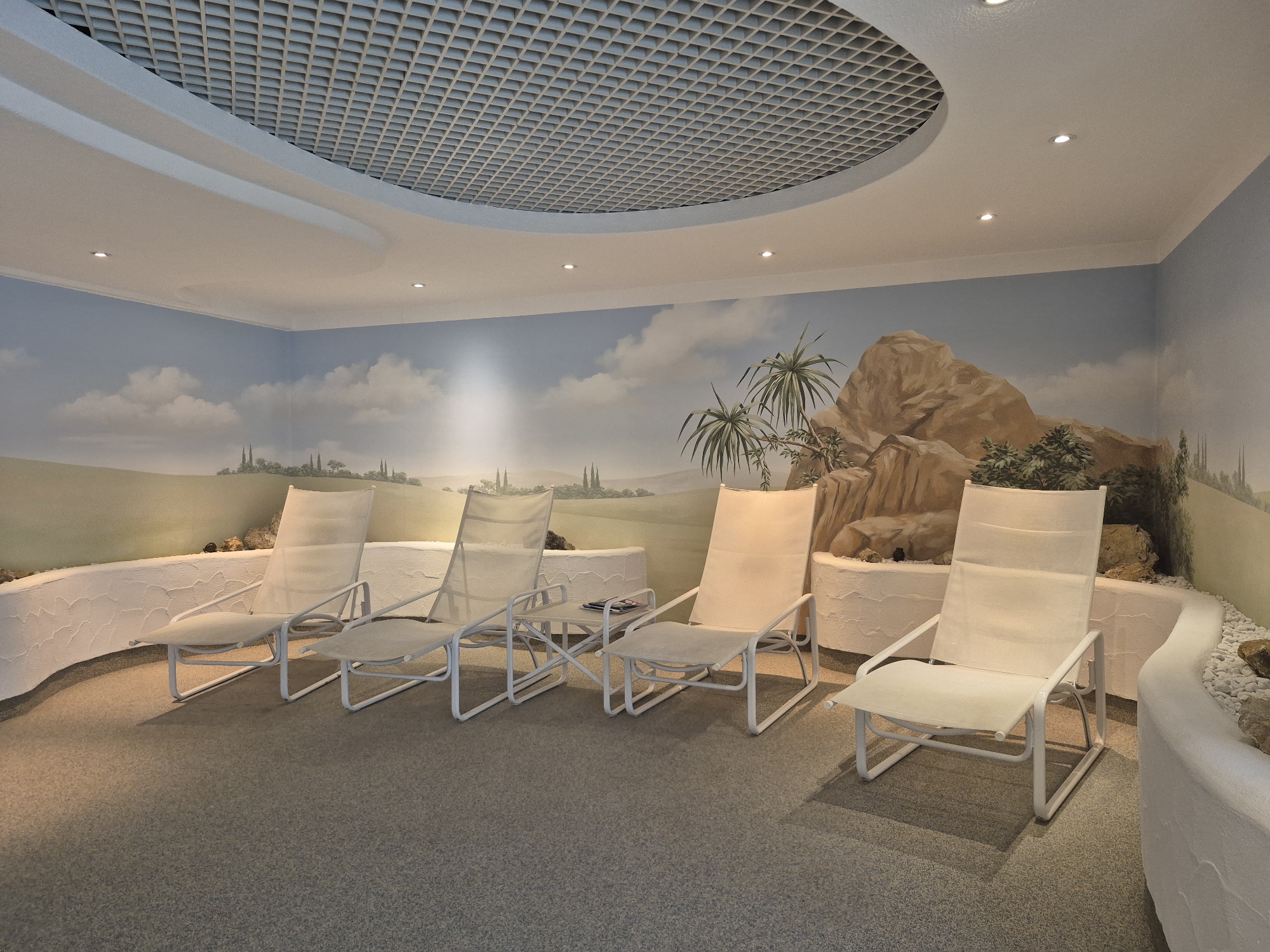 The image depicts a relaxation area, likely within a spa or wellness facility, situated in Schwäbisch Gmünd, Germany. The space is characterized by a serene ambiance, with a mural on the walls depicting a scenic landscape of rolling hills, distant trees, and rocky outcrops under a cloudy sky. Several white lounge chairs with light-colored fabric are arranged on a textured gray carpeted floor. A white, curved wall with a rough, stucco-like finish runs along the perimeter of the seating area, separating it from the mural. The ceiling features a distinctive, patterned grid design and recessed lighting. The overall impression is one of a tranquil indoor retreat designed for relaxation.