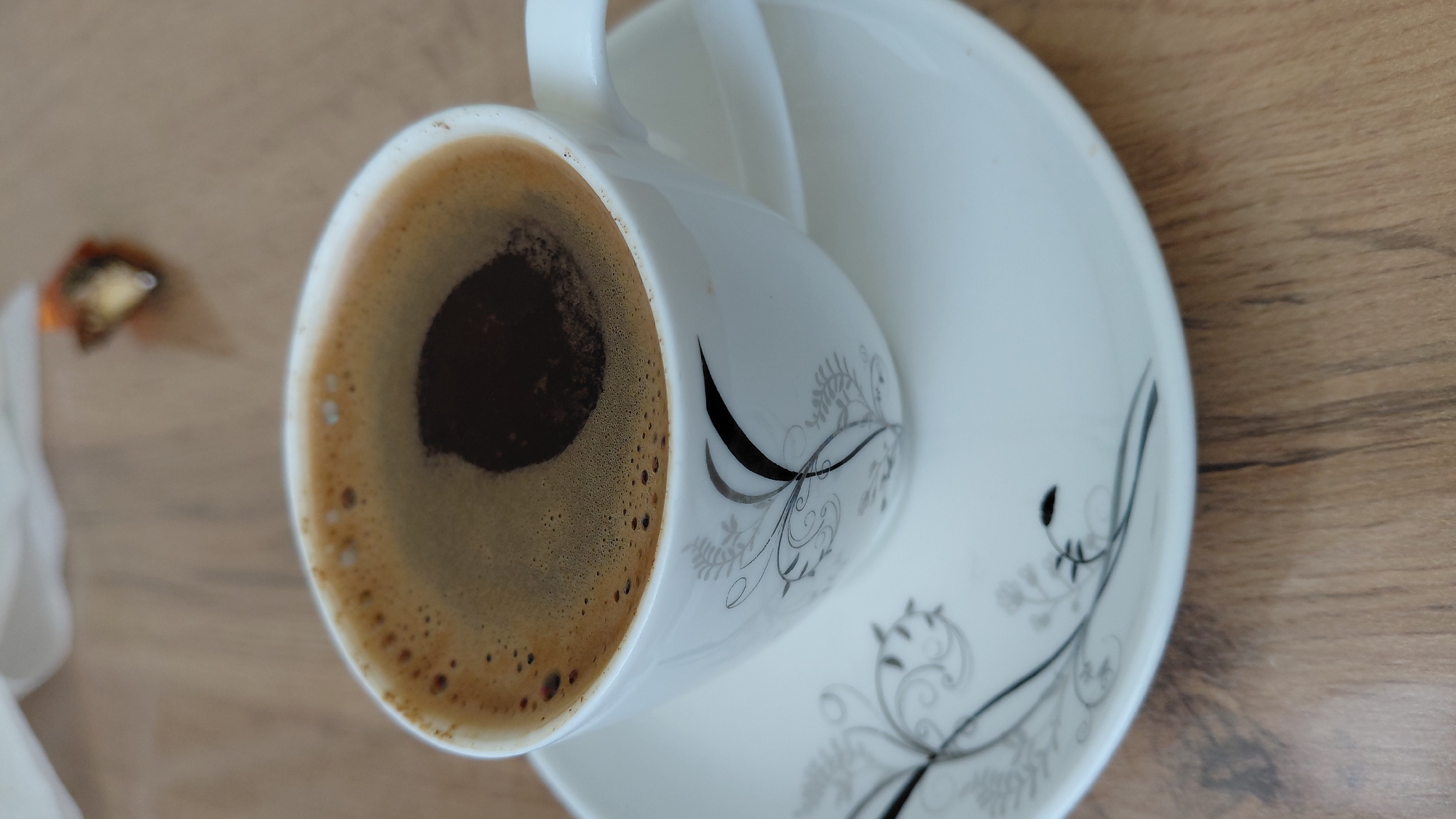 A close-up, high-angle shot shows a white porcelain coffee cup filled with dark, frothy coffee. The cup is adorned with a delicate black floral pattern and rests on a matching saucer. The coffee has a thick layer of crema with a darker pool in the center. The cup and saucer are placed on a light brown wooden surface, likely a table. In the background, a white napkin and a small, golden-wrapped candy are visible, slightly out of focus. This scene is situated in Abovyan, Armenia, suggesting a moment of quiet indulgence or a break during the day.