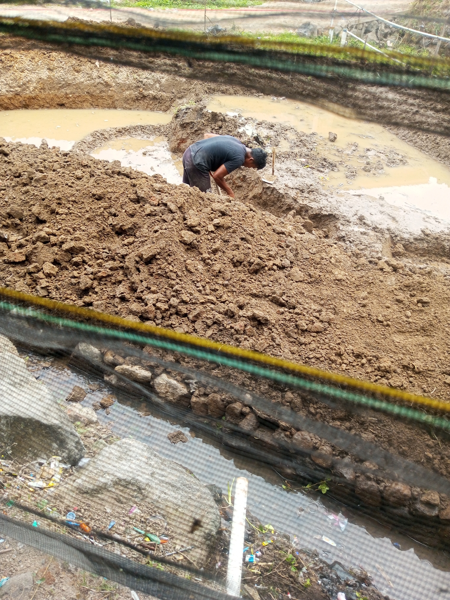 A person is digging in a muddy area in Kalibeber, Indonesia. The ground is a mixture of wet soil and dirt mounds. The person is wearing a dark t-shirt and shorts and is bent over, using a shovel. In the foreground, there is a net or barrier with green and yellow stripes, and beyond that, a ditch filled with water and debris, including plastic bottles and wrappers. The overall scene appears to be a construction or excavation site.