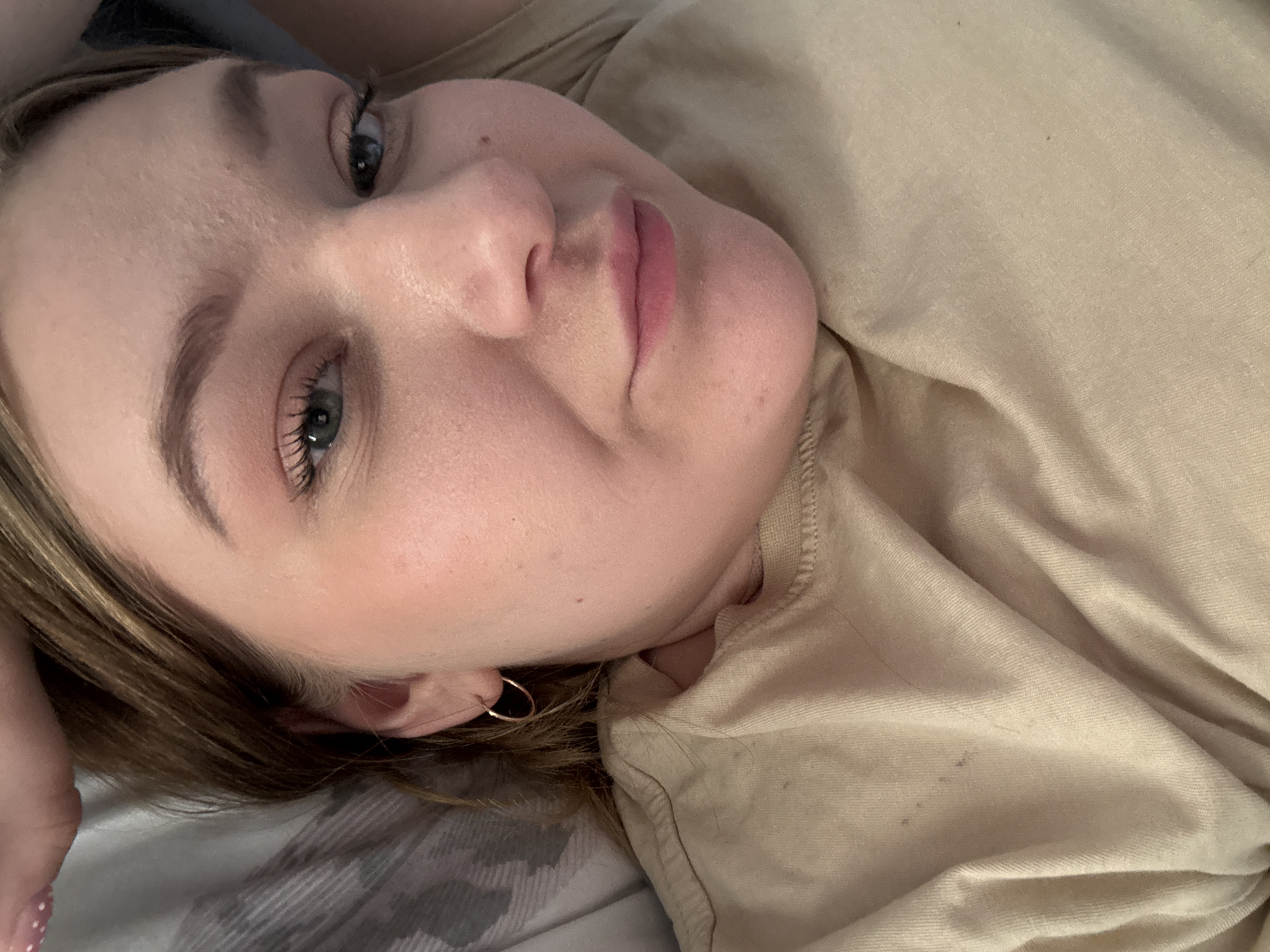 The scene depicts a close-up portrait of a single female subject, appearing adult, positioned in a reclining or lying posture. Her head rests on a gray and white patterned fabric, indicative of bedding. Her face, framed by medium brown hair with lighter highlights, is directed generally towards the camera, displaying blue-green eyes with mascara, defined eyebrows, and a subtle closed-mouth smile. The subject has fair skin, visible pores, light blush on the cheeks, and naturally pink lips. A small, dark mole is present on her right cheek/jawline. She wears a small, thin, gold-colored hoop earring in her visible left ear. Her attire consists of a light beige or tan crew-neck t-shirt, which exhibits minor wrinkles and faint dark specks on the left shoulder area. A hand, with light pink fingernails adorned with white polka dots, is partially visible in the upper left corner of the frame, near her head. The environment is an indoor residential space, likely a bedroom, situated in Chaikovsky, Russia.