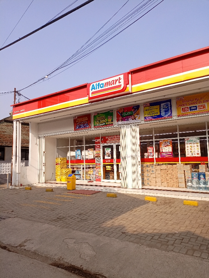 The image shows an Alfamart convenience store in Katapang, Indonesia. The store is located at the coordinates -6.984, 107.554. The store is a one-story brick building with a red and yellow awning. The storefront is made of glass, and there are several signs hanging from the awning. There are several rows of packaged goods and drinks on shelves inside the store, some of which are visible through the windows. There is a yellow trash can in front of the store, on the paved parking lot, and there are three yellow bollards to the left and right of the entrance. The street in front of the store is paved and there is a small, gray strip of sidewalk in front of the store. In the distance, a dark-colored building is visible. The sky is blue and there are power lines overhead. There are no people visible in the image.