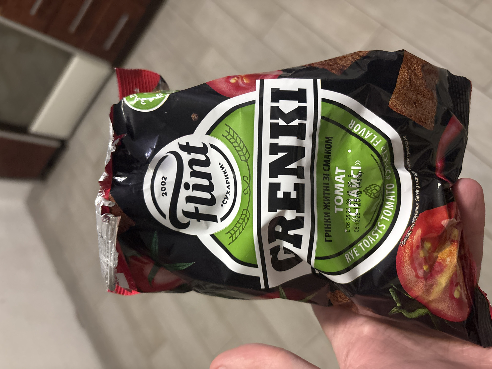 The image shows a close-up of a hand holding a package of "Flint" brand rye toasts with tomato flavor. The packaging is predominantly black with a green and white design. The brand name "Flint" is prominently displayed in large white lettering. Below that, in smaller text, are the words "СУХАРИКИ" which translates to "croutons" or "rusks" in Russian. Further down, the product name "GRENKI" is in large bold letters, with "ГРІНКИ ЖИТНІ ЗІ СМАКОМ" (Rye croutons with flavor) above it and "ТОМАТ" (Tomato) below. The phrase "RYE TOASTS TOMATO FLAVOR" is also visible on the packaging. The image is taken indoors, with a blurred background that suggests an indoor environment, possibly a kitchen or pantry, with wooden furniture visible in the distance. There are no indications of a specific city or location within the image itself. A slice of tomato is depicted on the packaging, and a few of the rye toasts are also visible, appearing as small, dark brown squares. The packaging is slightly opened at the top.