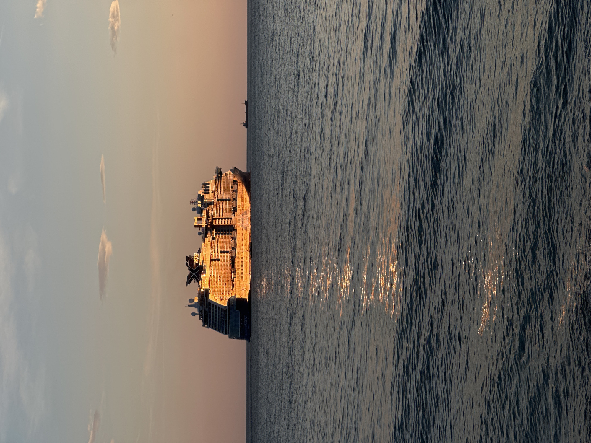 A cruise ship, named "Celebrity Ascent," is sailing in the sea off the coast of Šušanj, Montenegro. The ship is illuminated by the warm light of the setting or rising sun. The sea surface is ruffled by wind, with reflections of sunlight creating shimmering patterns. The sky above is a pale color with some light clouds.