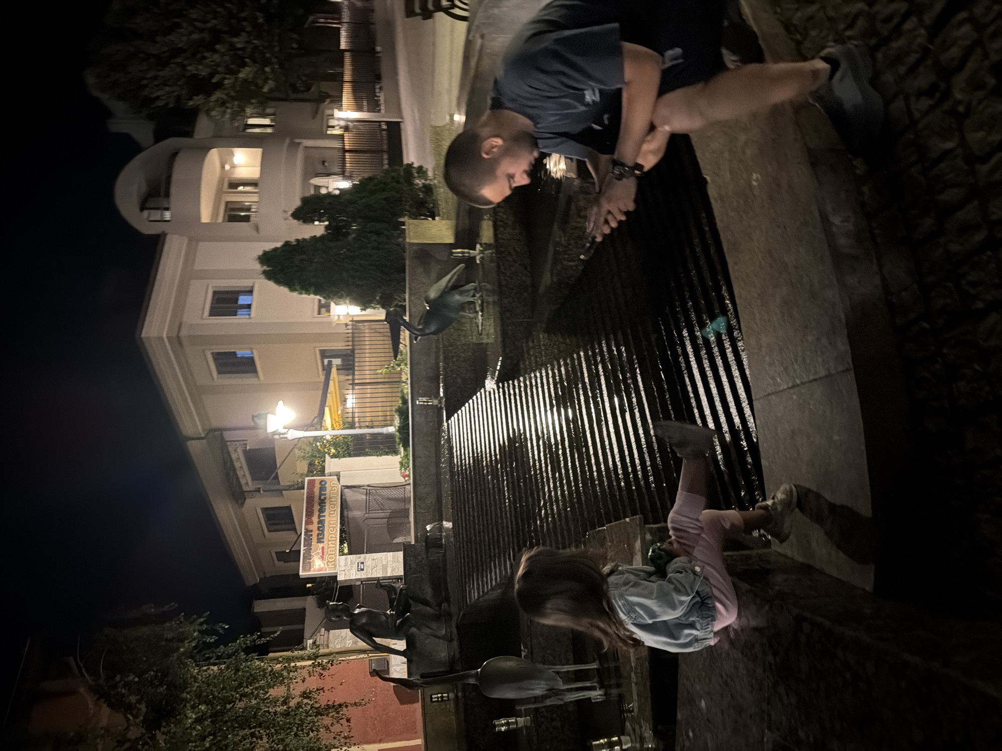 At night in Burgas, Bulgaria, a young girl and a man are seated at the edge of a tiered fountain. The girl, facing away from the camera and towards the fountain's water, wears a light blue denim jacket, pink pants, and light-colored sneakers. She holds a green object in her lap. The man, to the right and also facing the fountain, wears a dark t-shirt and dark shorts, and is looking down at his mobile phone. His legs are crossed. The fountain has dark, stepped surfaces where water cascades down, illuminated by lights. In the background, across a dark street, a multi-story building with illuminated windows and balconies stands. To the left of the building, a sign with Cyrillic text is visible, indicating "IZDATELSTVO" (Publishing House) and "KOPIREN TSENTUR" (Copy Center). Statues of human figures and birds are integrated into the fountain's design. The surrounding area appears to be a public space with trees and streetlights.