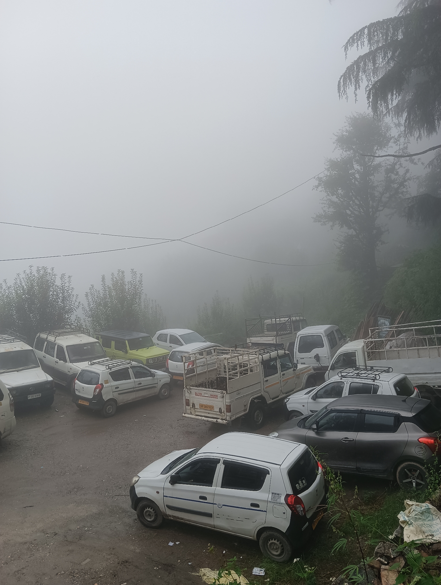 The image depicts a parking lot filled with various vehicles, including cars, SUVs, and trucks, situated on a dirt and gravel surface. The scene is enveloped in thick fog, which obscures the background and creates a hazy atmosphere. Trees and vegetation are visible through the fog, suggesting an outdoor setting, likely in a hilly or mountainous region. The vehicles appear to be parked haphazardly, with some close to one another. The overall impression is one of a quiet, possibly secluded location, characterized by the dense fog and the presence of numerous parked vehicles. The location is identified as Siumsa, India.
