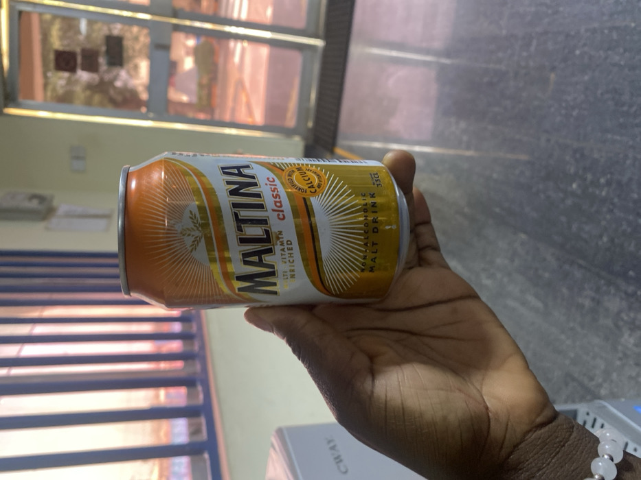 A dark-skinned right hand, adorned with a light-colored beaded bracelet on the wrist, holds an orange, white, and gold 33cl Maltina Classic non-alcoholic malt drink can. The can's label indicates it is "MULTI VITAMIN ENRICHED" and "FORTIFIED WITH CALCIUM AND VITAMINS." The scene is indoors, located in Kaduna South, Nigeria. In the background to the left, a window with vertical blue blinds allows warm, pinkish-orange light to enter. Partially visible to the far left is a white appliance labeled "CWAY." To the right, a glass door reflects ambient light and shows indistinct outdoor elements beyond. The flooring appears to be a light-colored, reflective surface.