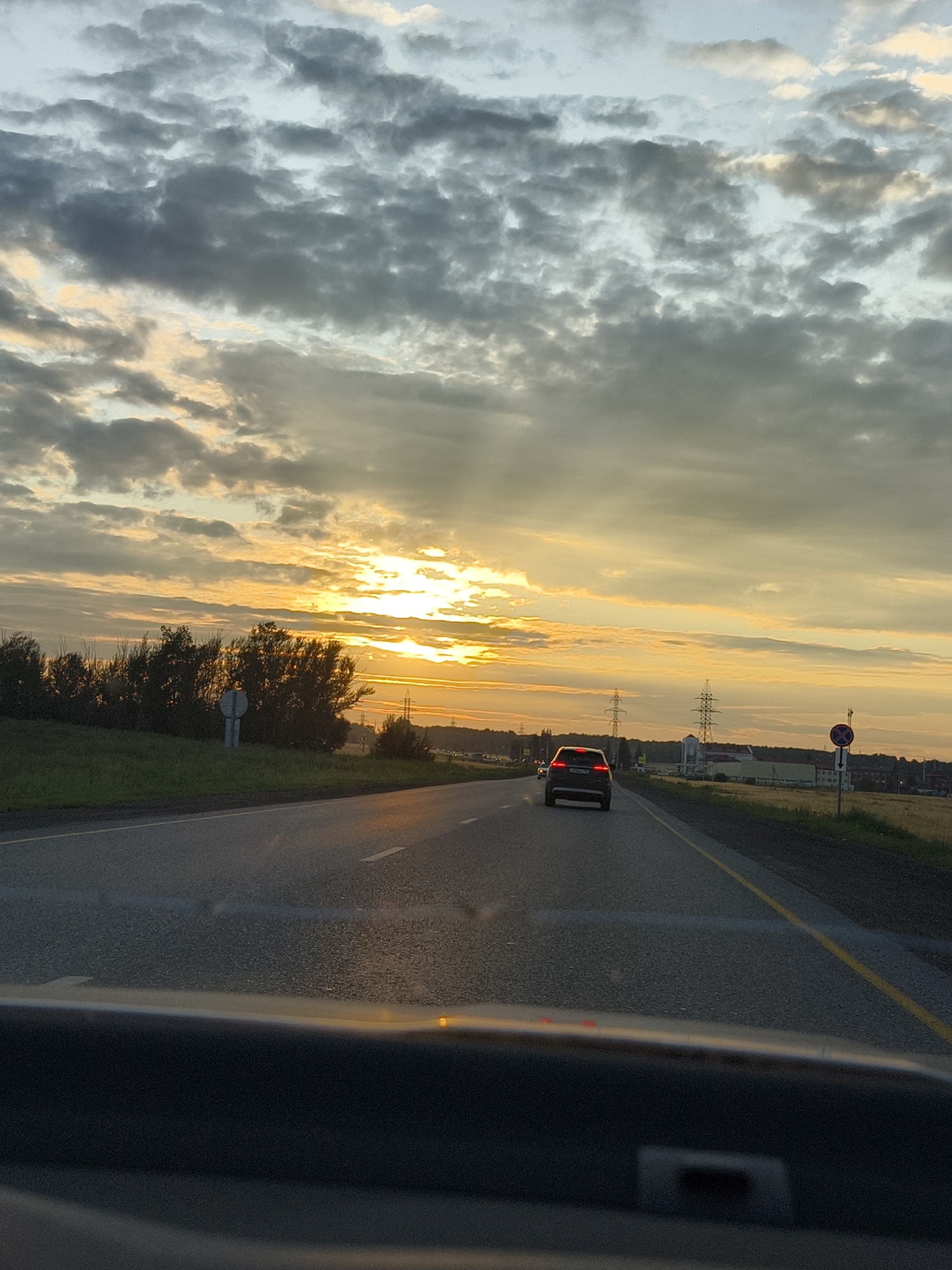 This image depicts a sunset scene on a highway, likely in or near the city of Kuraskovo, Russia. The sky is filled with dramatic clouds, with rays of sunlight breaking through, illuminating the scene with a warm, golden glow. A dark-colored sedan is traveling on the right lane of the two-lane highway. The road ahead curves slightly to the left. On the right side of the road, there are fields and power lines. On the left side, there are trees and vegetation. The foreground shows the dashboard of a vehicle, indicating the photo was taken from inside a car. The time of day is dusk, given the sunset lighting.