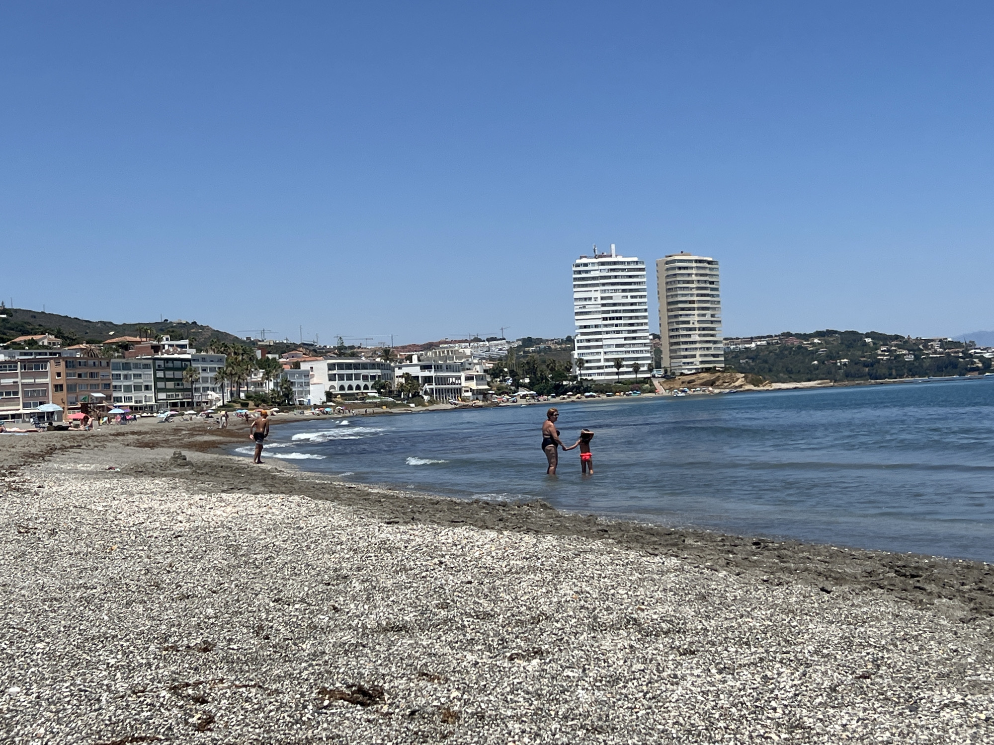 The image depicts a beach scene in San Roque, Spain, under a clear blue sky. In the foreground, a pebble beach stretches towards the calm, blue sea. Several people are visible on the beach and in the water. A woman and a child are standing in the shallow water, facing each other. Further down the beach, a lone person stands near the water's edge. In the middle ground, a line of buildings, including apartment complexes and hotels, forms the urban backdrop to the beach. Two tall, white apartment buildings with balconies are prominent features in the cityscape. The coastline extends into the distance, with hills covered in greenery and more scattered buildings. The overall impression is of a sunny, populated beach destination.
