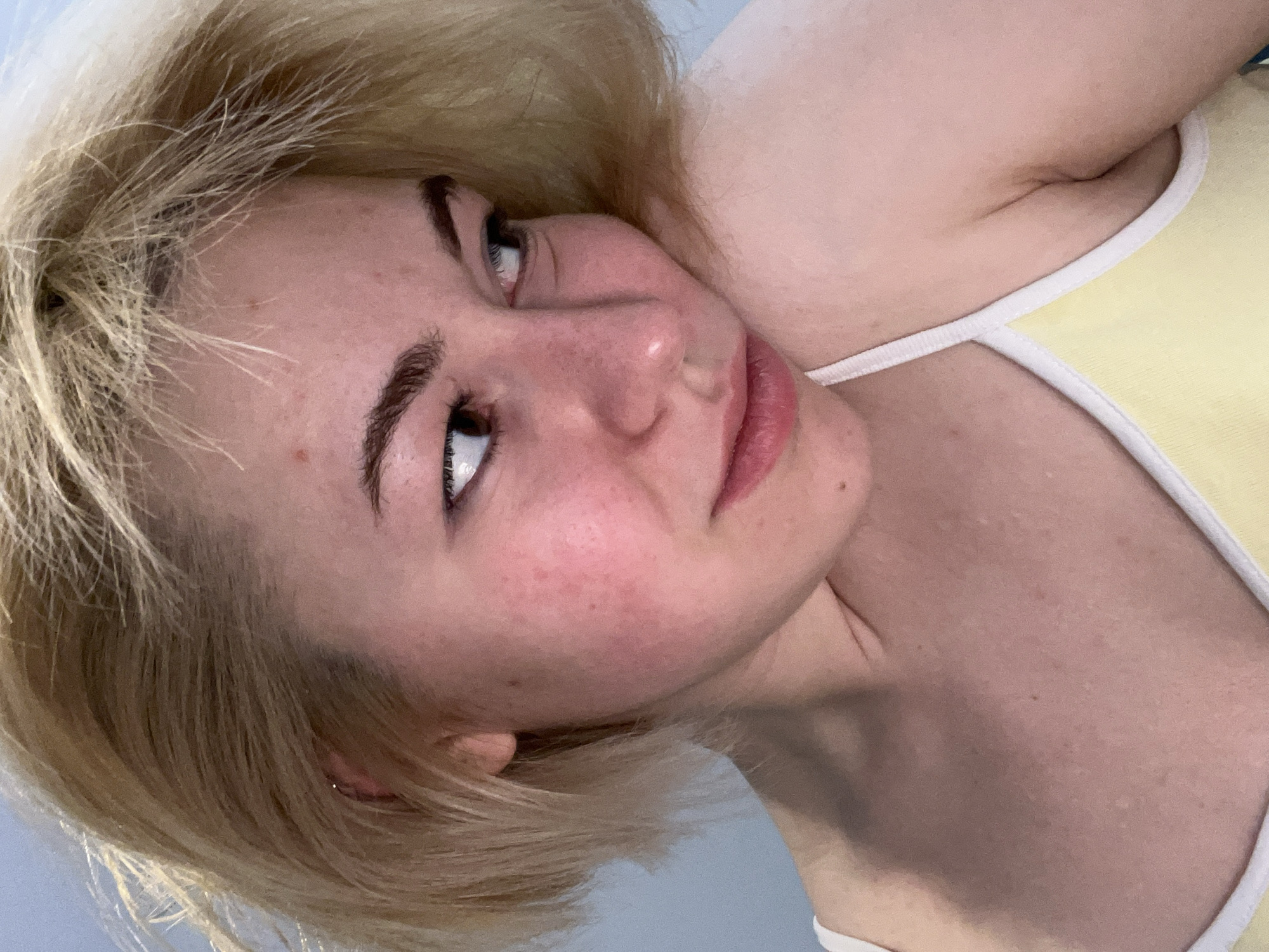A close-up portrait, taken at a tilted angle, features a young woman with light skin and blonde, shoulder-length hair. Her head is resting on a surface, and she gazes upwards and slightly to her left with dark, well-defined eyes. Her eyebrows are dark and prominent, and her full lips have a natural pink hue. A subtle redness is visible on her cheeks. She is wearing a light yellow top or garment with white trim along the neckline and straps. The background is a plain, light blue or grey, suggesting an indoor setting. Her expression appears calm and contemplative. No specific time of day, weather, or visible text can be discerned from the image, which originates from Boreti, Montenegro.