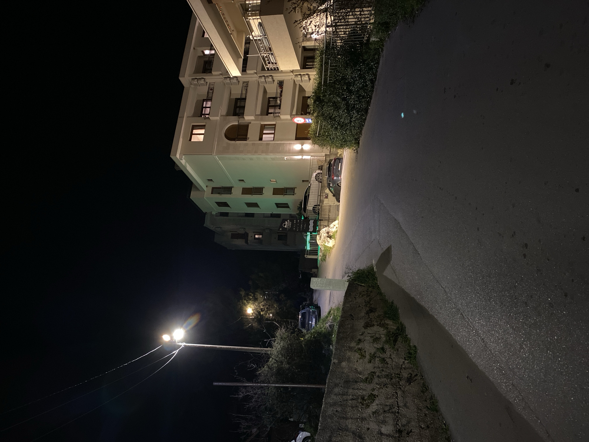 The image shows a street scene at night in Budva, Montenegro. A tall, light-colored building with multiple floors and balconies dominates the left side of the frame. The building is illuminated with warm interior lights visible through some of the windows. Adjacent to the building is a dark patch of greenery and foliage. A street with asphalt pavement runs along the right side of the frame. The street is dimly lit. Street lights are visible in the distance on the left, casting a glow. The environment is dark, with minimal ambient light. The image captures a quiet urban scene in Budva at night. There are cars parked along the street and near the building. The building has an entrance with a sign, indicating a possible business. The foreground on the right side is a stone wall covered with foliage.