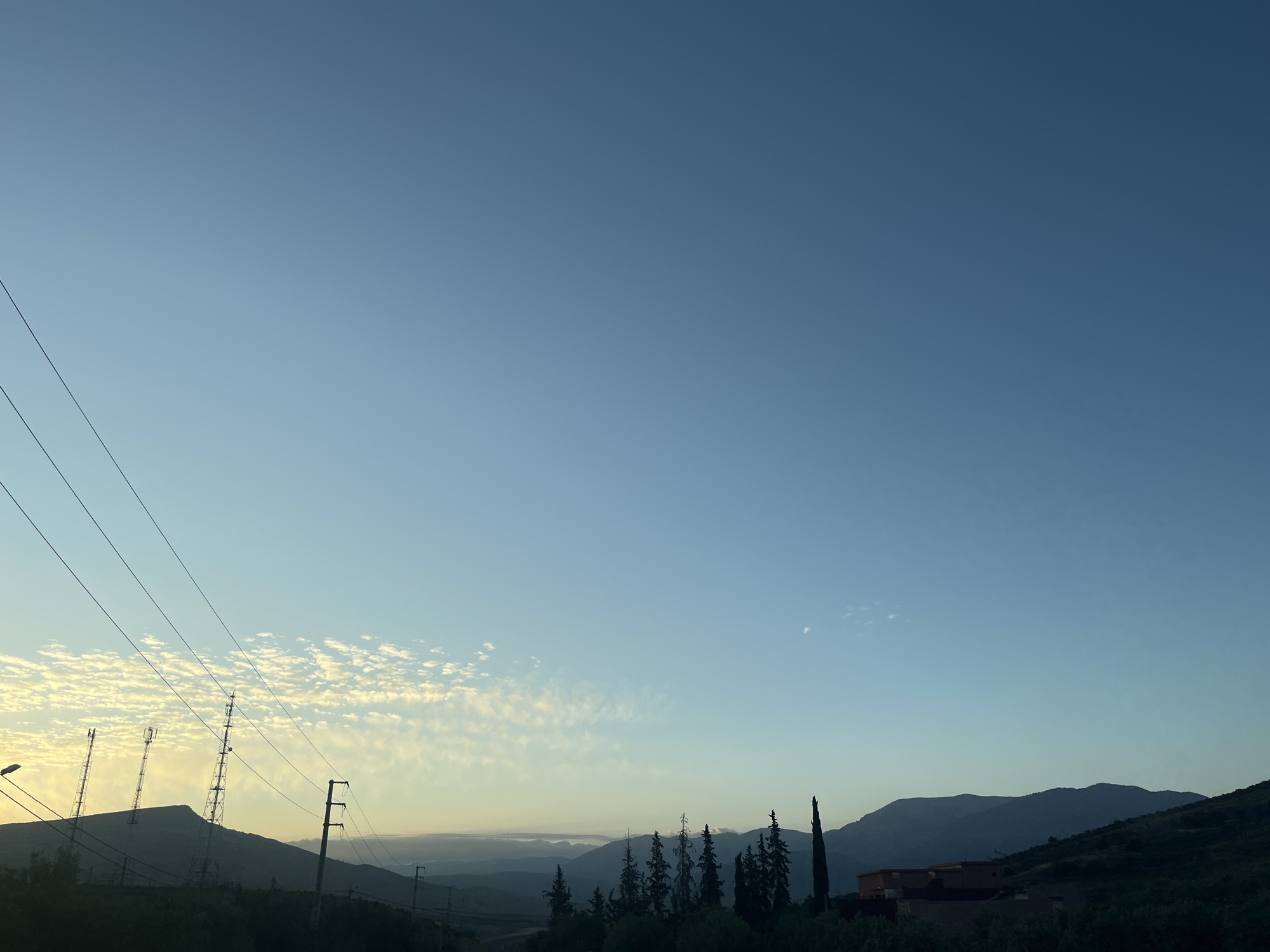 The photograph captures a landscape near Ouaouizert, Morocco, at dawn or dusk. The sky transitions from a light blue near the horizon to a deeper blue overhead. Altocumulus clouds are scattered in the distance.
Mountains form the horizon, their silhouettes varying in height and shape. In the foreground, there are evergreen trees and a building. Power lines stretch across the left side of the frame. Several communication towers are visible on a hill.