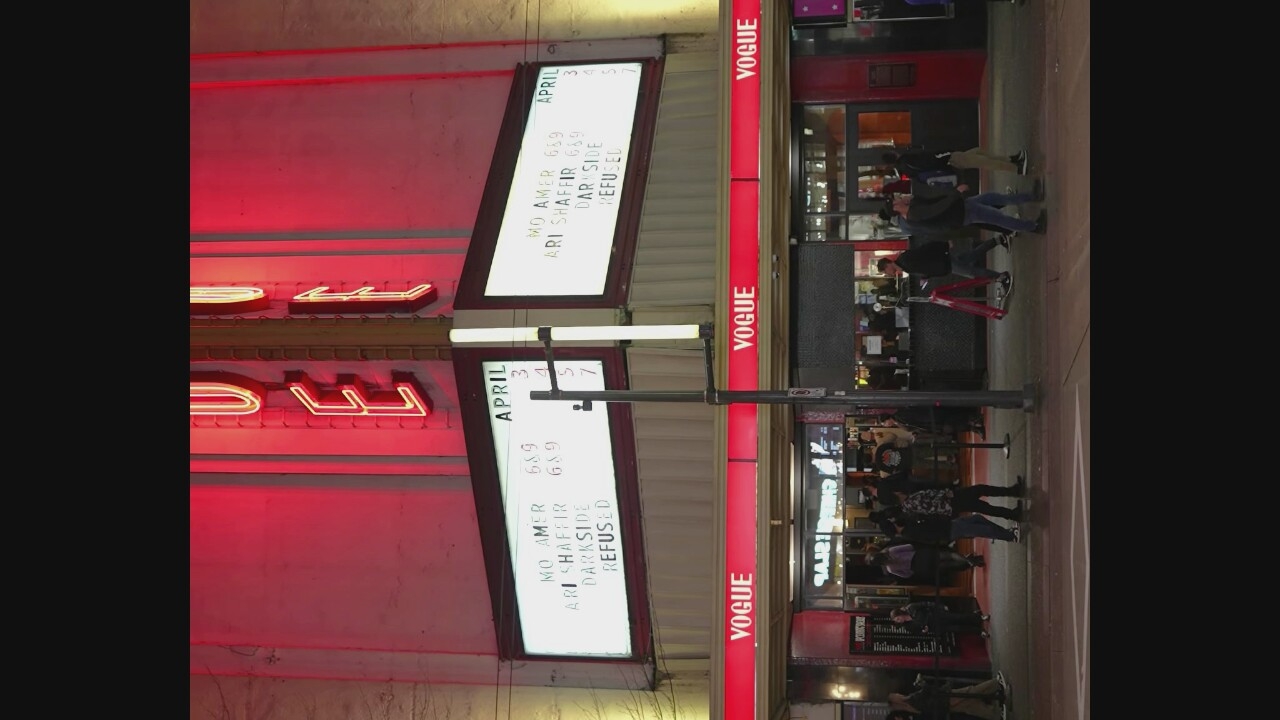 The image shows the exterior of the Vogue Theatre in Vancouver, Canada, at night. The theatre's marquee displays upcoming events. The neon sign above the marquee spells out "VOGUE" in red and yellow lights. The marquee itself is illuminated white and shows several lines of text, including "APRIL," "MO AMER," "ARI SHAFFIR," "DARK SIDE," and "REFUSED," along with corresponding numerical times.
The entrance to the theatre is visible at street level, with red signage reading "VOGUE" above each entrance. People are seen walking on the sidewalk in front of the theatre. Some individuals are standing near the entrance, possibly in line. A red A-frame sign is also visible on the sidewalk. The street in front of the theatre is paved, with white lines indicating a crosswalk. The overall scene depicts a lively evening at a popular entertainment venue.