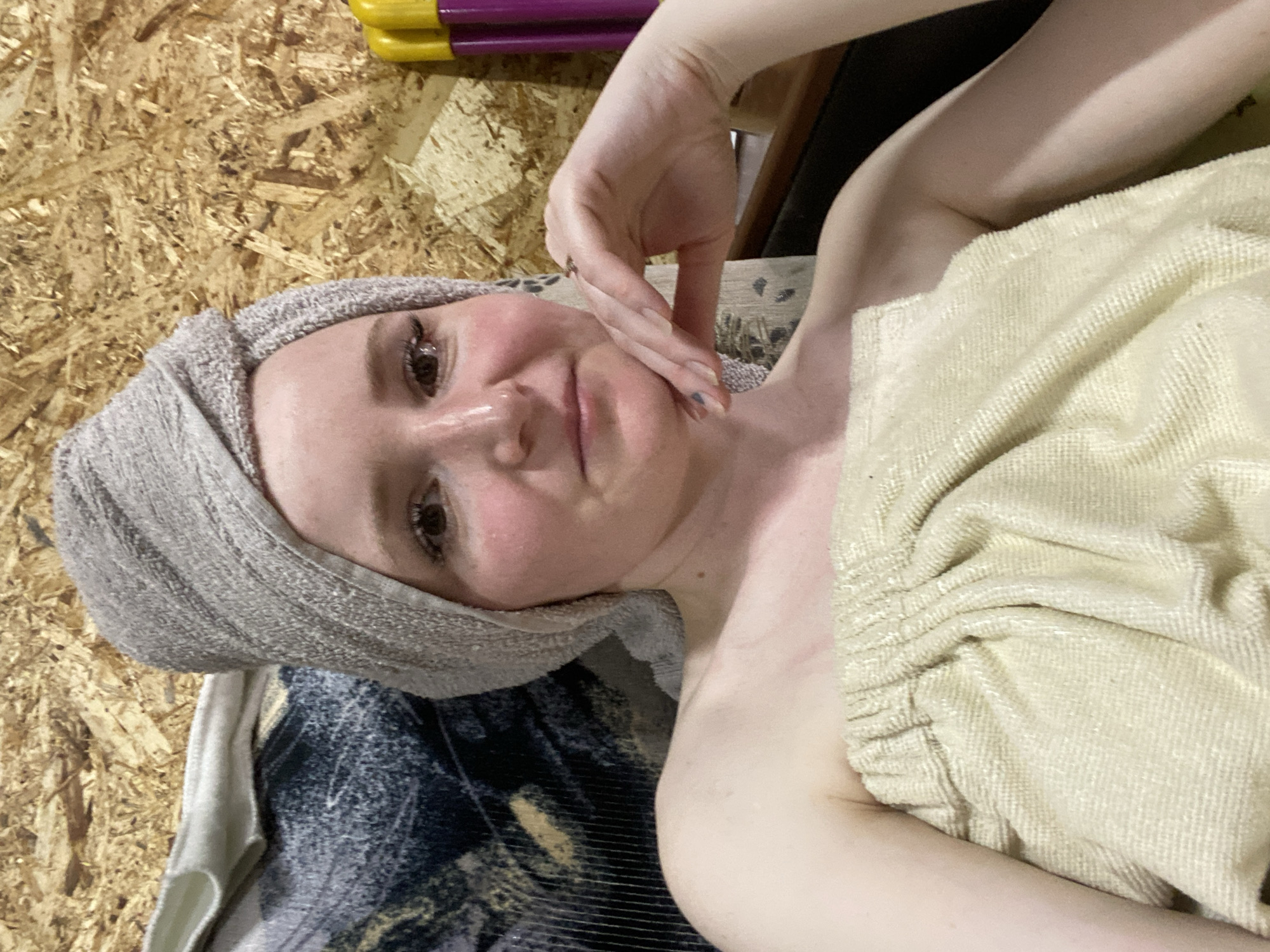 A young adult female is depicted indoors in Chaikovsky, Russia, looking directly at the camera with a slight, closed-mouth smile. Her light brown hair is wrapped in a light grey/beige towel, turban-style, and her body is draped in a light yellow/beige towel, covering her chest and shoulders. She exhibits a flushed complexion on her cheeks and wears a small nose ring in her right nostril. Her left hand, with partially visible dark nail polish on the thumb and index finger and a small ring on the ring finger, rests gently under her chin. The background consists primarily of an Oriented Strand Board (OSB) wall. To the subject's left, a patterned dark blue and gold fabric is partially visible, along with a lighter beige fabric. To her right, two upright cylindrical objects, one yellow and one purple, resembling spray cans or bottles, are positioned on a dark horizontal surface.