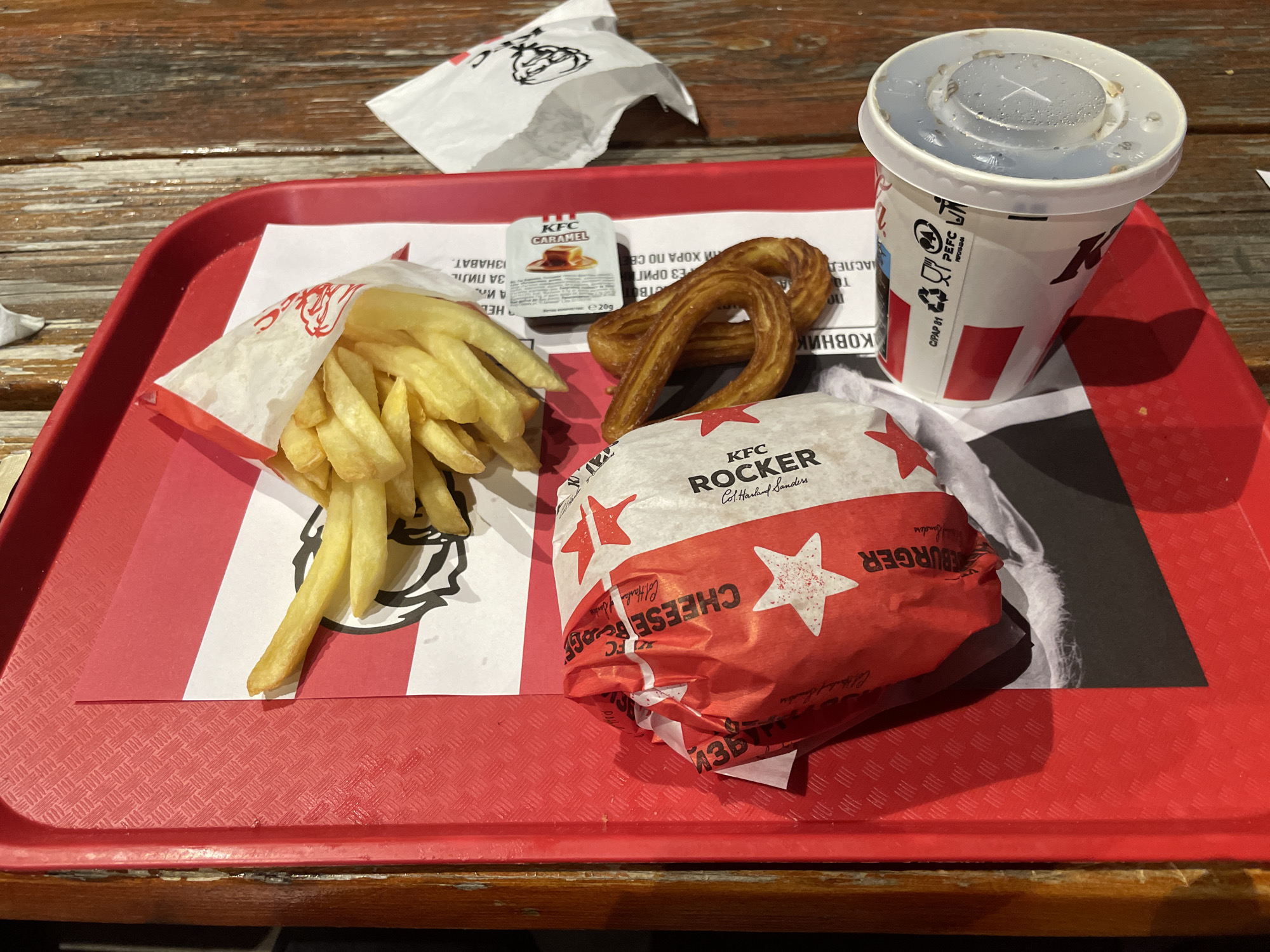 A red, textured plastic tray, lined with a white paper sheet featuring red stripes and the KFC logo, holds a fast-food meal on a weathered wooden table. The meal includes a serving of golden-yellow french fries contained within a white paper pouch bearing KFC branding and Cyrillic text. Adjacent to the fries are two golden-brown, twisted churros. A "KFC ROCKER" burger, wrapped in red paper adorned with white stars and displaying Cyrillic text "ЧИЙЗБУРГЕР" (Cheeseburger), rests beside the churros. A white KFC branded paper cup with red stripes and a clear plastic lid, showing condensation and splatter, contains a beverage. A small white packet of "KFC Caramel" sauce, labeled "е 20g", is positioned near the churros. A crumpled white paper item with the KFC logo is visible in the background. The Cyrillic script on the packaging elements indicates the scene is located in a Bulgarian-speaking region, specifically identified as Sofia, Bulgaria.