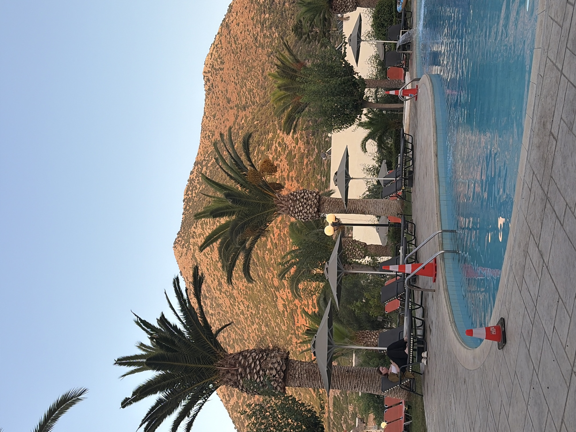A wide-angle, eye-level shot captures an outdoor swimming pool area in Vlichada, Greece. The foreground features the turquoise water of a curved swimming pool, bordered by a paved patio. Two orange and white traffic cones are placed on the patio, one near the pool's edge and another by a set of metal pool stairs. Several empty lounge chairs and umbrellas are visible, some on the patio and others on a grassy area in the background. A woman sits on a lounge chair, looking down at something in her lap, while several palm trees are interspersed throughout the scene. Behind the pool area, a steep, sun-drenched mountain covered in scrub vegetation dominates the background, its rocky surface illuminated by the warm light of what appears to be late afternoon or early evening. The sky is a clear, pale blue.