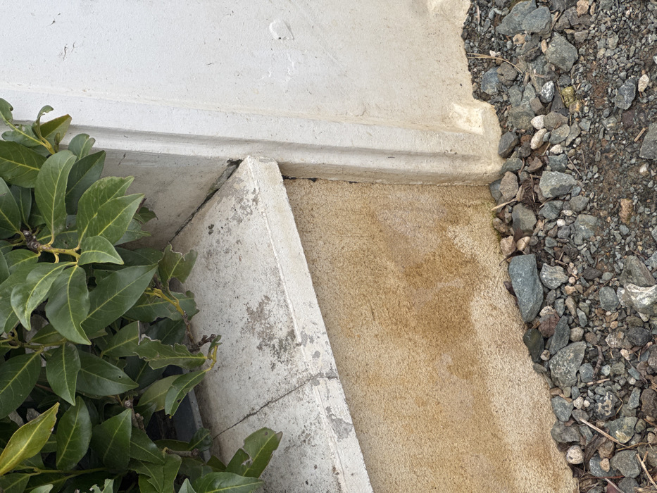 This image depicts a static exterior scene in Echzell, Germany, focusing on the lower section of a building or wall and adjacent ground. The foreground consists of irregularly shaped, multi-colored gravel in shades of grey and brown, interspersed with small organic debris such as dried leaves and twigs. Directly above the gravel is a textured, reddish-brown foundation wall, exhibiting vertical streaks and patches of darker discoloration, consistent with moisture staining. Resting on top of this foundation wall is a lighter-colored, possibly concrete, coping stone with visible surface dirt, minor chipping, and a distinct crack extending horizontally across its top surface. To the immediate right, a vertical corner of an off-white painted or stucco wall features a prominent vertical architectural relief. Partially obscuring the upper left corner of the coping and extending out of frame is a dense green shrub or hedge with broad, ovate leaves, some of which show minor insect damage along their edges. No individuals or animate subjects are present.