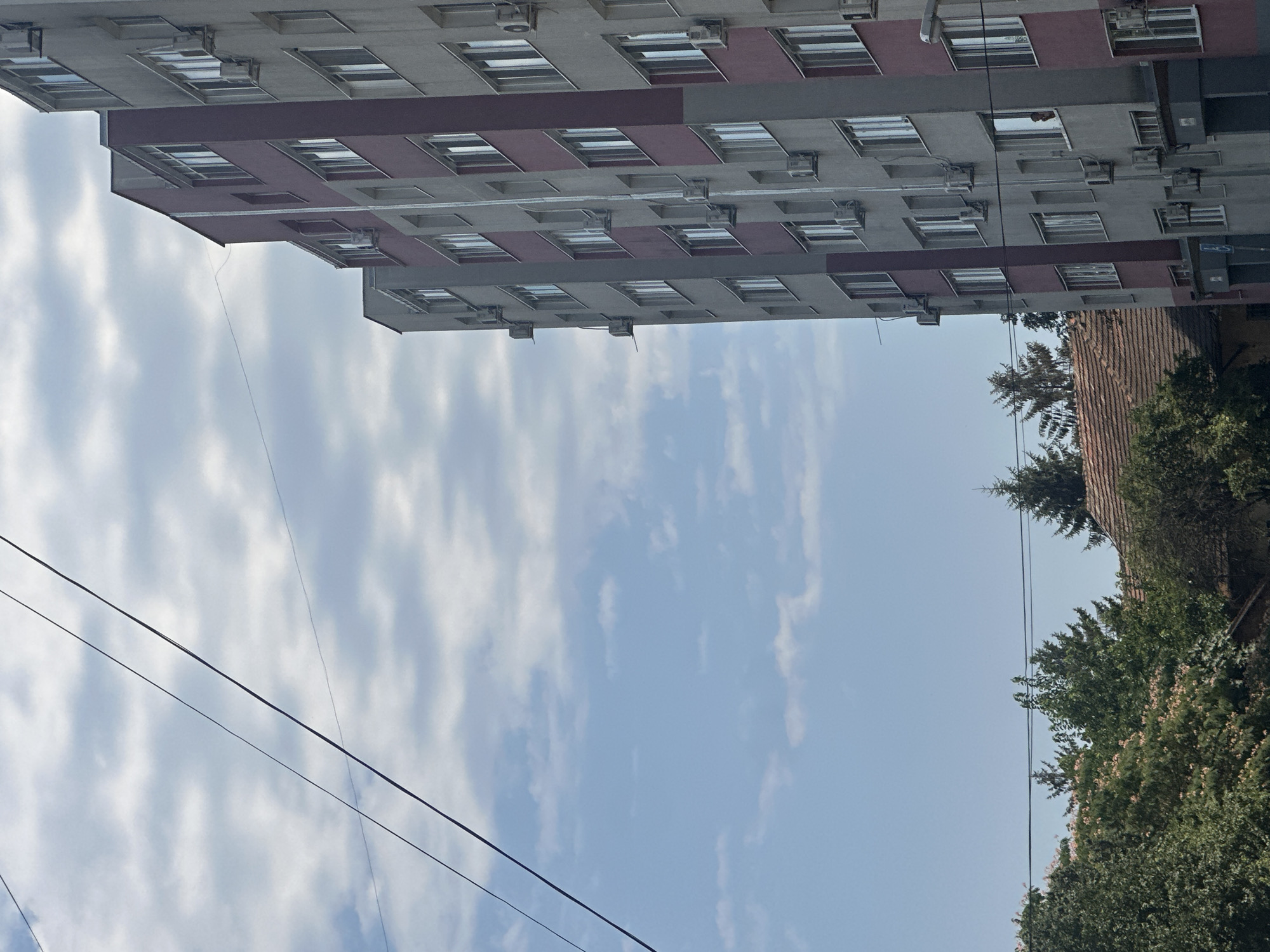 The image shows a side view of a multi-story apartment building with a light gray facade and a maroon-colored vertical strip. Numerous windows with white frames are visible, and air conditioning units are mounted beneath many of them. Power lines crisscross the sky. In the lower left, there are trees and the roof of a smaller building. The sky is blue with scattered white clouds.