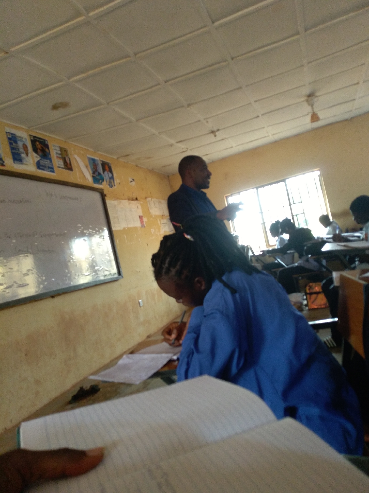 A medium shot captures a classroom scene in Jos, Nigeria, with a teacher gesturing towards a whiteboard as students are seated at desks. The classroom appears somewhat aged, with a stained yellow wall and a grid-patterned ceiling. A whiteboard on the left displays some handwritten text, mostly illegible due to reflections and distance.
In the foreground, a student's back is visible, wearing a blue garment. Their hands are engaged in writing on a notebook, with another notebook lying open in front of them. Further into the background, the teacher, a dark-skinned man with a bald head, is standing and talking, facing away from the camera but turned towards the students. Several other students are seated at desks, some appearing to be writing or looking forward.
A large window with metal bars is visible in the background, letting in bright daylight. The overall impression is of a typical classroom setting where learning is taking place, albeit in a modestly equipped environment. The presence of posters on the wall and the general state of the room suggest a long-standing educational institution.