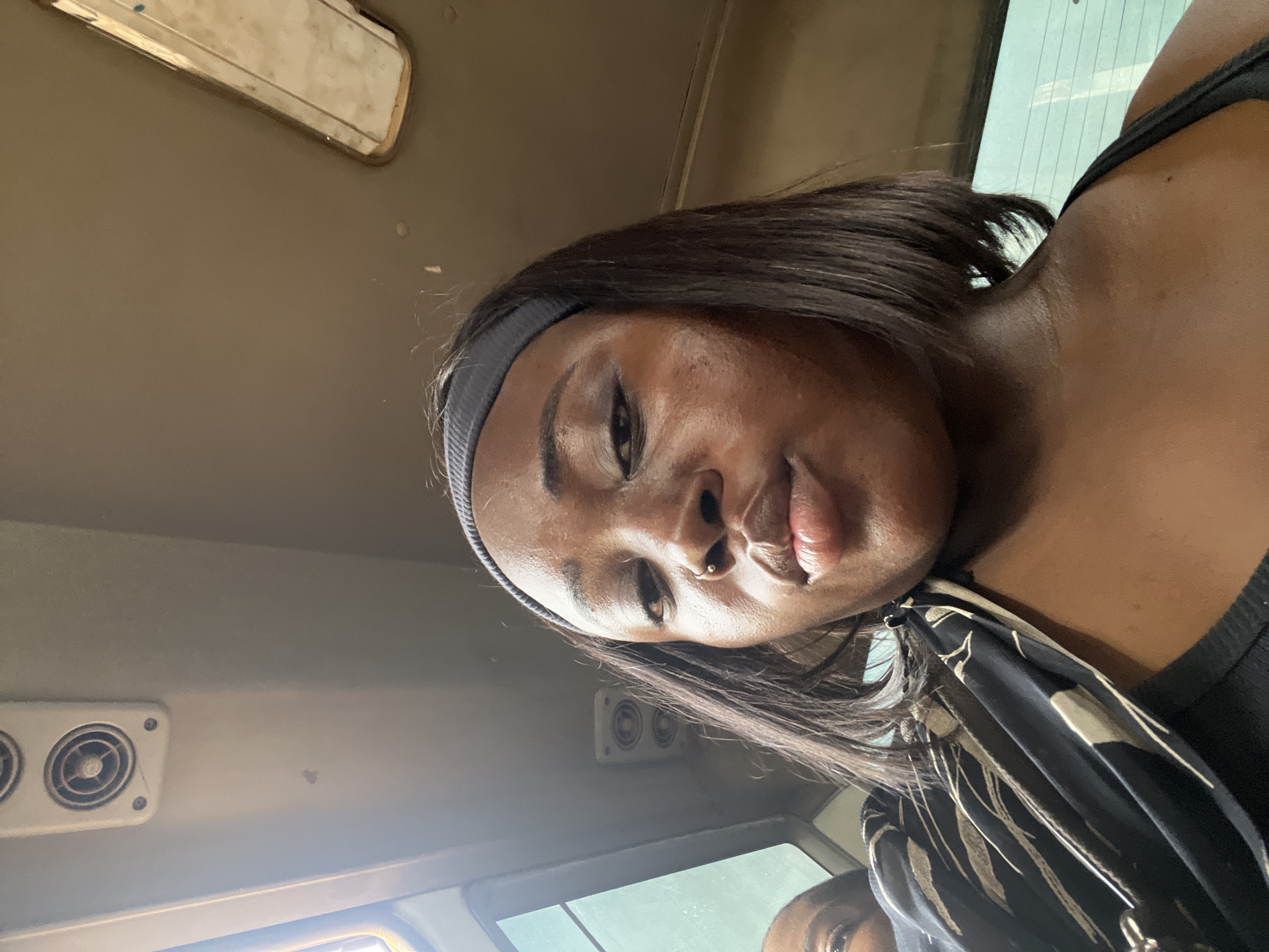 This is a selfie taken by a young Black woman inside a vehicle, likely a bus. She is wearing a grey headband and a black top. She has a nose ring. Her hair is dark and straight, falling around her face. The background shows the interior of the vehicle, including air vents and a window. The lighting suggests it is daytime, and the reflection in the window indicates an outdoor scene. The woman's expression is neutral, and she is looking directly at the camera. A second person's face is partially visible in the lower right corner of the frame. The scene appears to be within Kugbo, Nigeria.