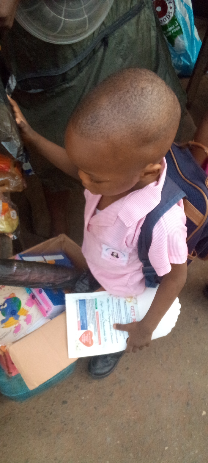 A young individual, likely a boy, with a shaved head and dark skin, is depicted from an elevated perspective in Nnewi, Nigeria. He wears a pink, collared, short-sleeved, ribbed shirt with a dark blue backpack and black shoes. A small white rectangular badge featuring a cartoon-like image and text is pinned to the left side of his shirt. He is holding a white document titled "CITY 202" with a red heart graphic. His left arm extends towards the left, near a clear plastic bag containing an orange-yellow item. A brown cardboard box filled with school supplies, including a colorful notebook, is positioned below his arm. The background features a dark green fabric or tarp, a white and blue plastic bag partially visible with the word "FOODS," and a clear, ribbed plastic object. The ground surface is a light brown, appearing to be concrete or packed dirt.